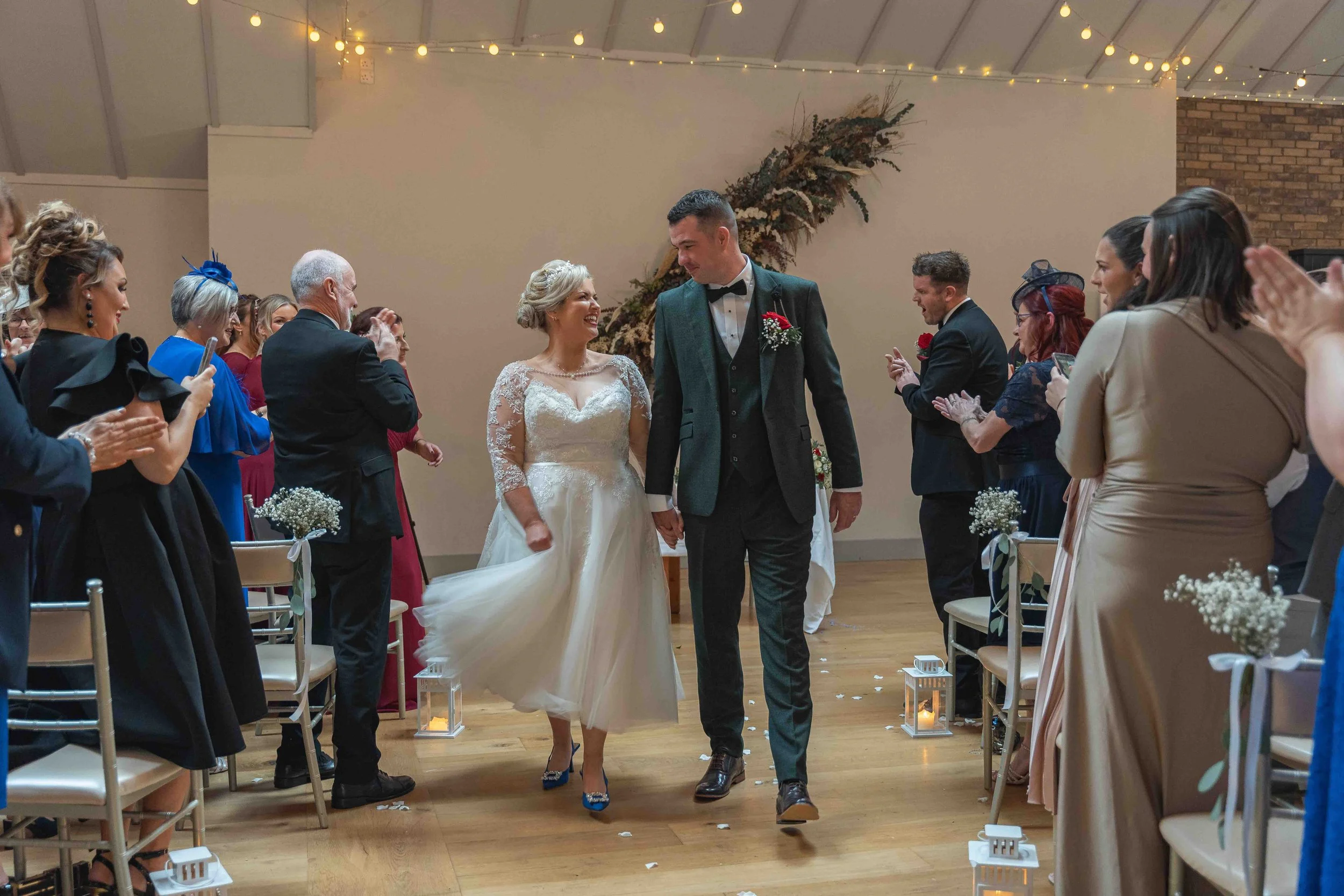 Bride and groom walking down the aisle at their wedding ceremony, surrounded by seated guests clapping and smiling in a decorated indoor venue.