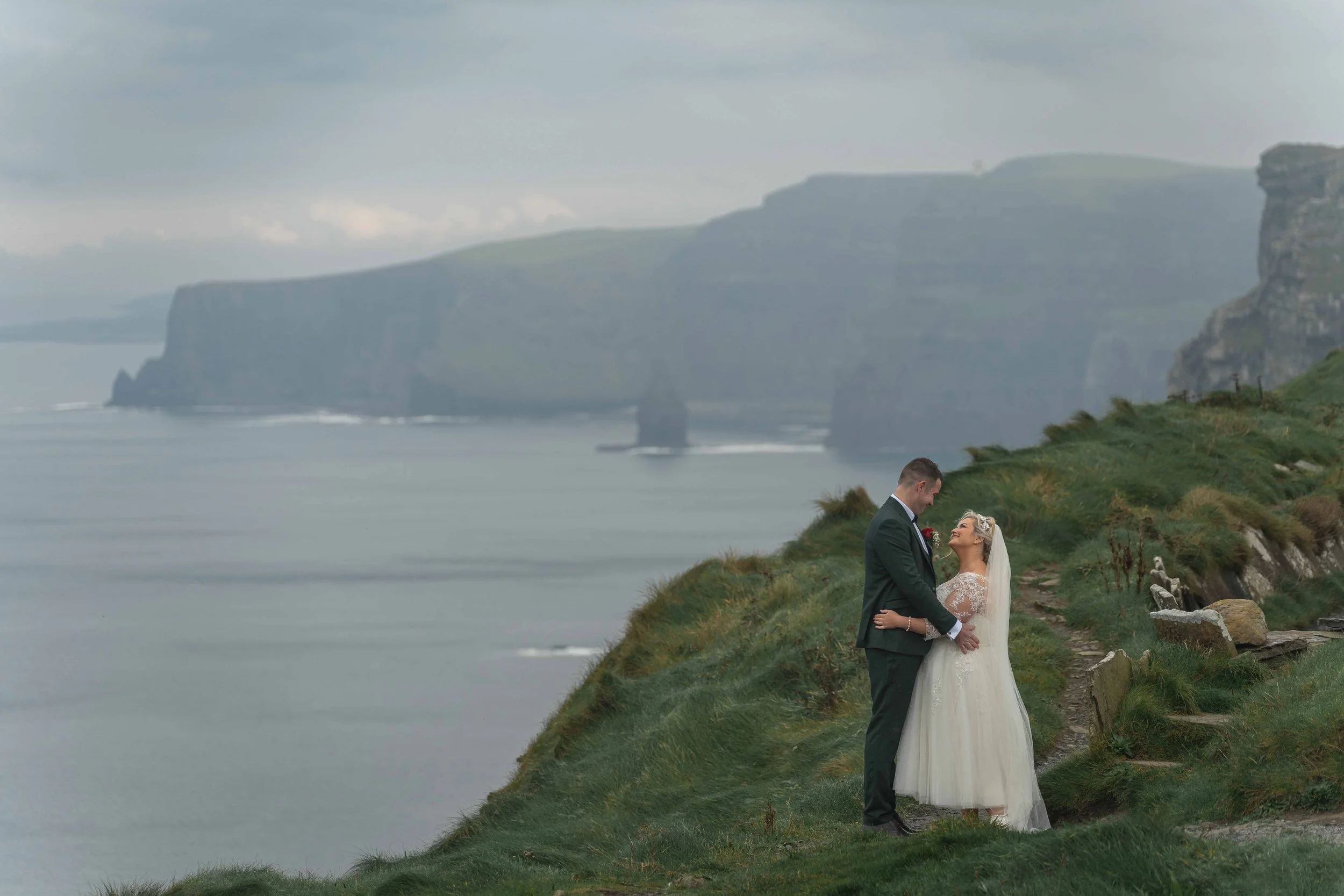 A bride and groom standing together on a grassy cliffside overlooking the ocean, with rocky cliffs in the background, during a wedding celebration.