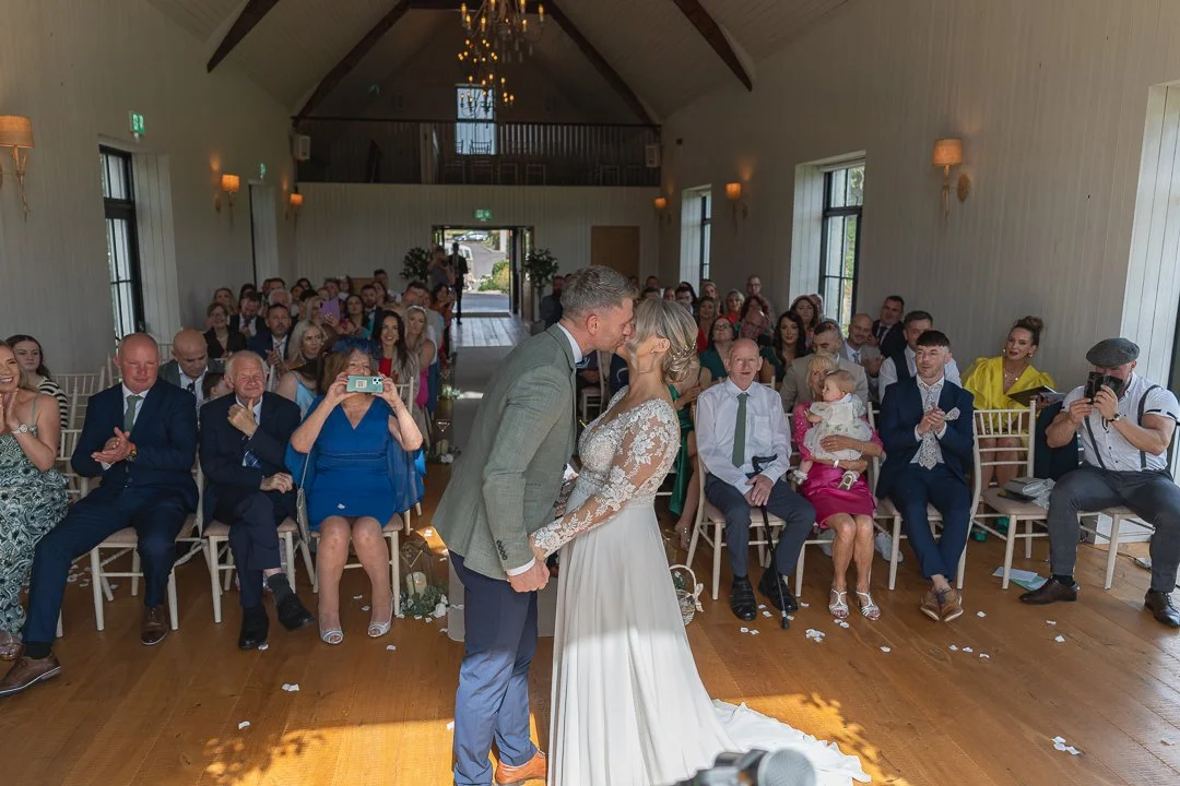 Bride and groom share a kiss during their wedding ceremony inside a bright, rustic hall, with guests seated and watching.