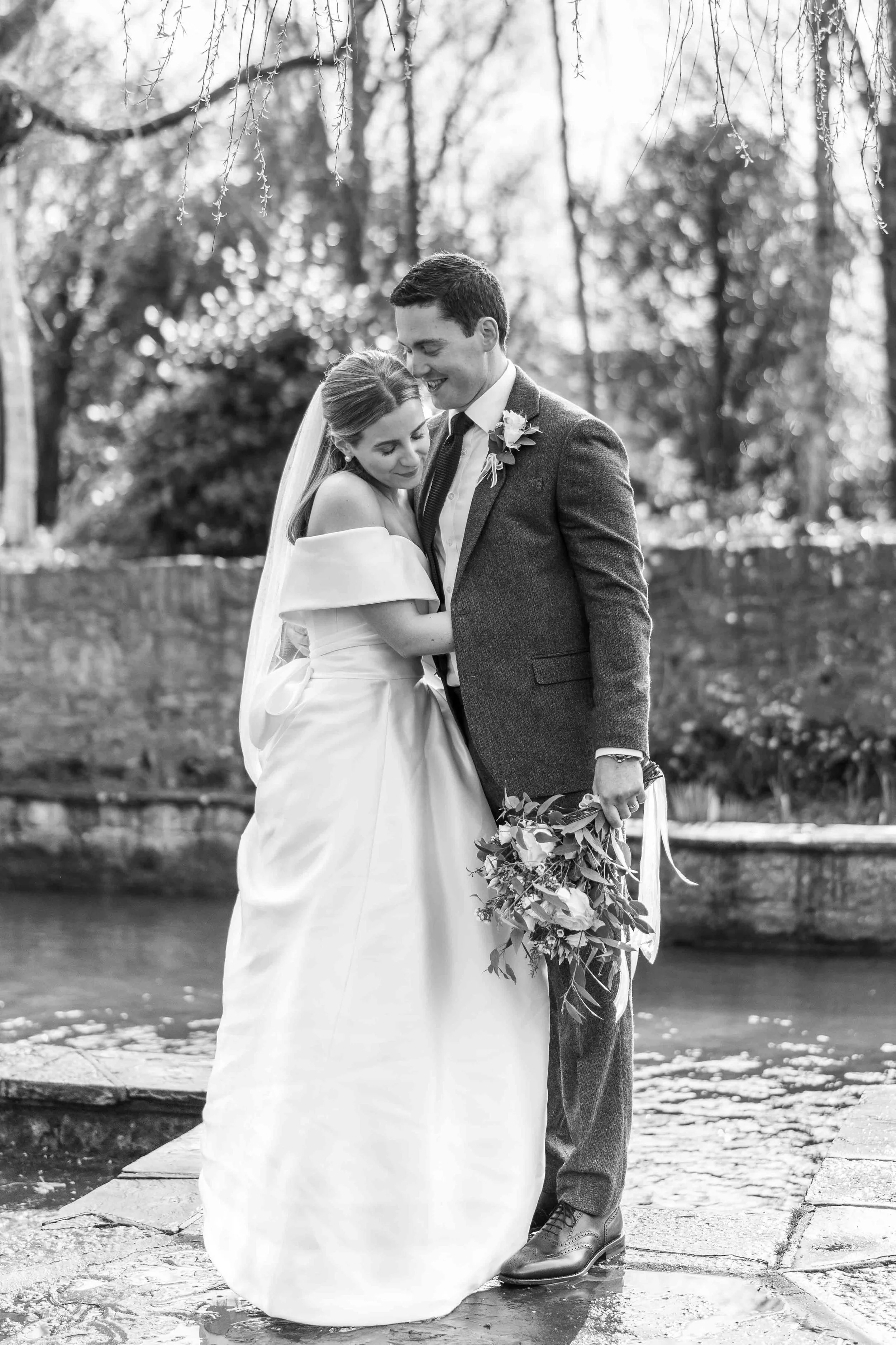 Black and white photo of a bride and groom embracing outdoors, standing on stone pavement near a pond, with trees in the background.
