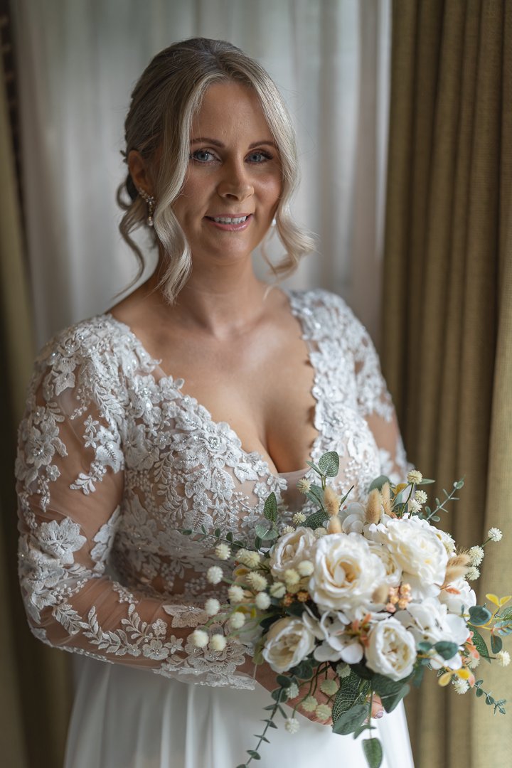 A bride holding a bouquet of white and cream flowers with green foliage, wearing a lace wedding dress with floral embroidery, posing near a window with curtains.