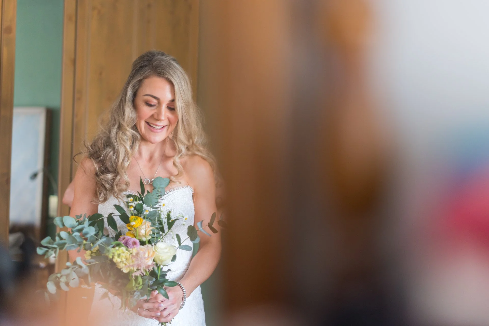 A smiling woman in a white dress holding a bouquet of flowers, standing indoors with wooden walls.