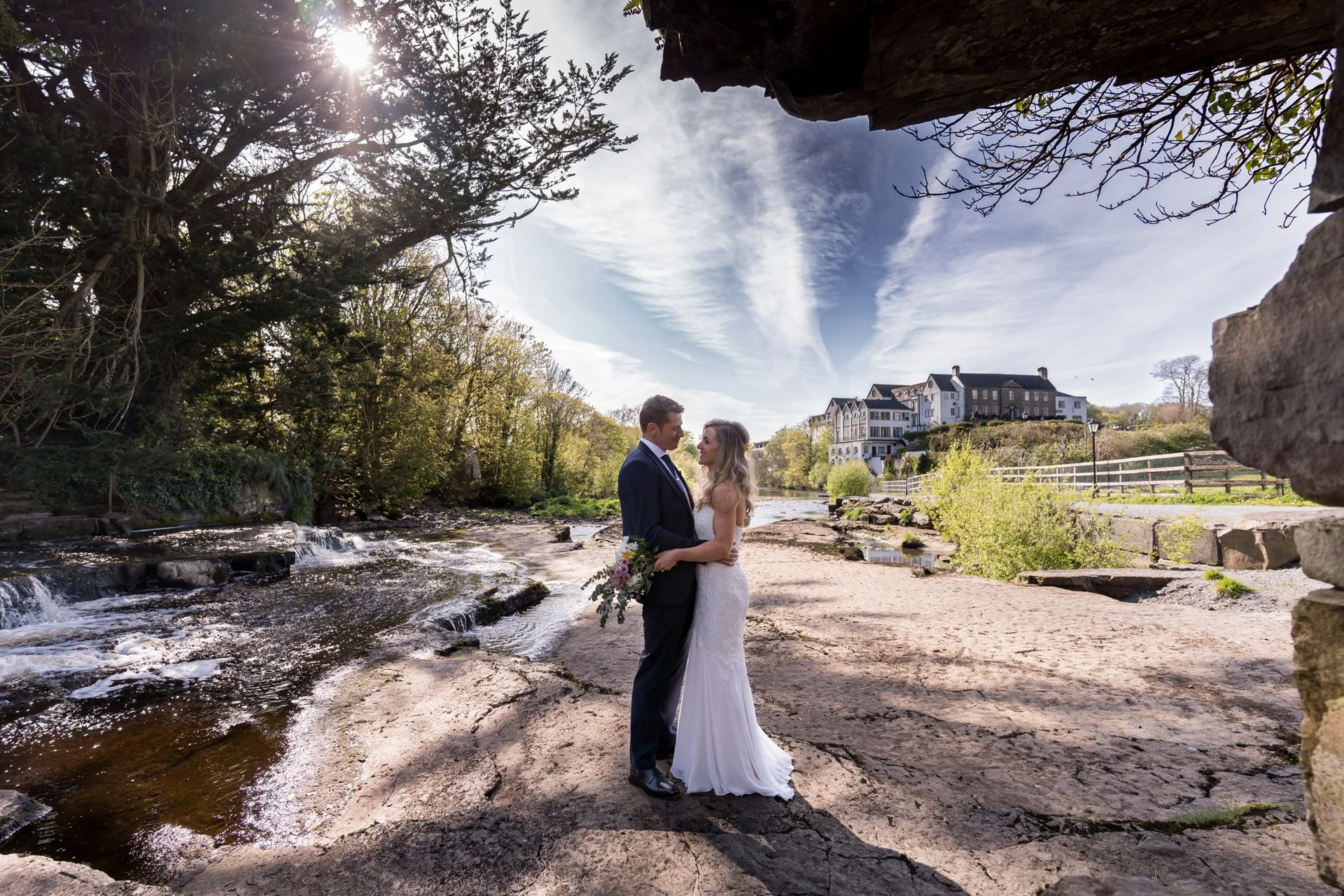 A bride and groom are standing on a rocky riverbank, facing each other and holding hands, with a scenic background of trees, a flowing river, a large house or hotel on a hill, and a partly cloudy sky.