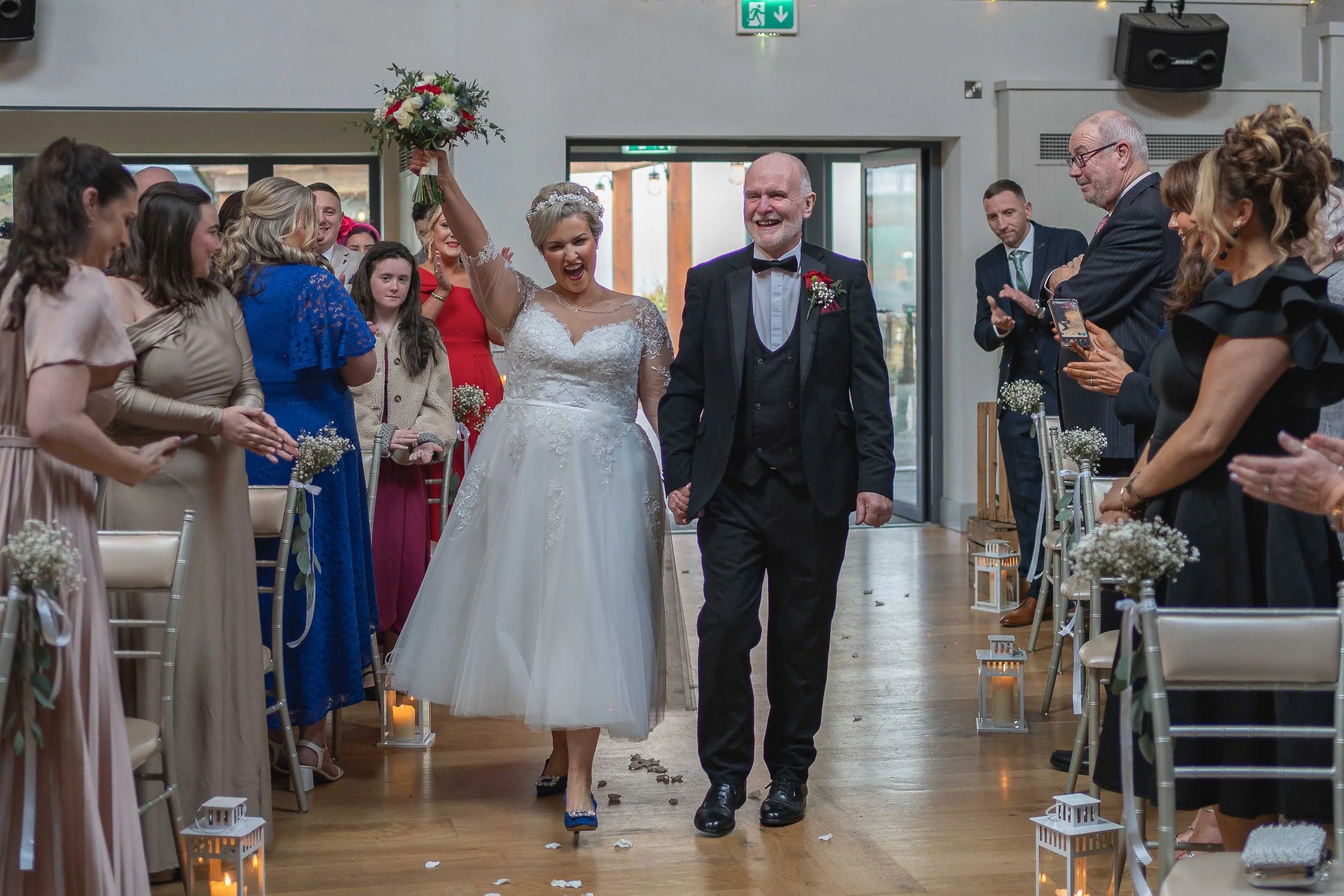 A bride and an older man, possibly her father, walking down the aisle at a wedding ceremony. The bride is holding a bouquet high in celebration and smiling. Guests on both sides of the aisle are clapping and taking photos. The indoor venue is decorat