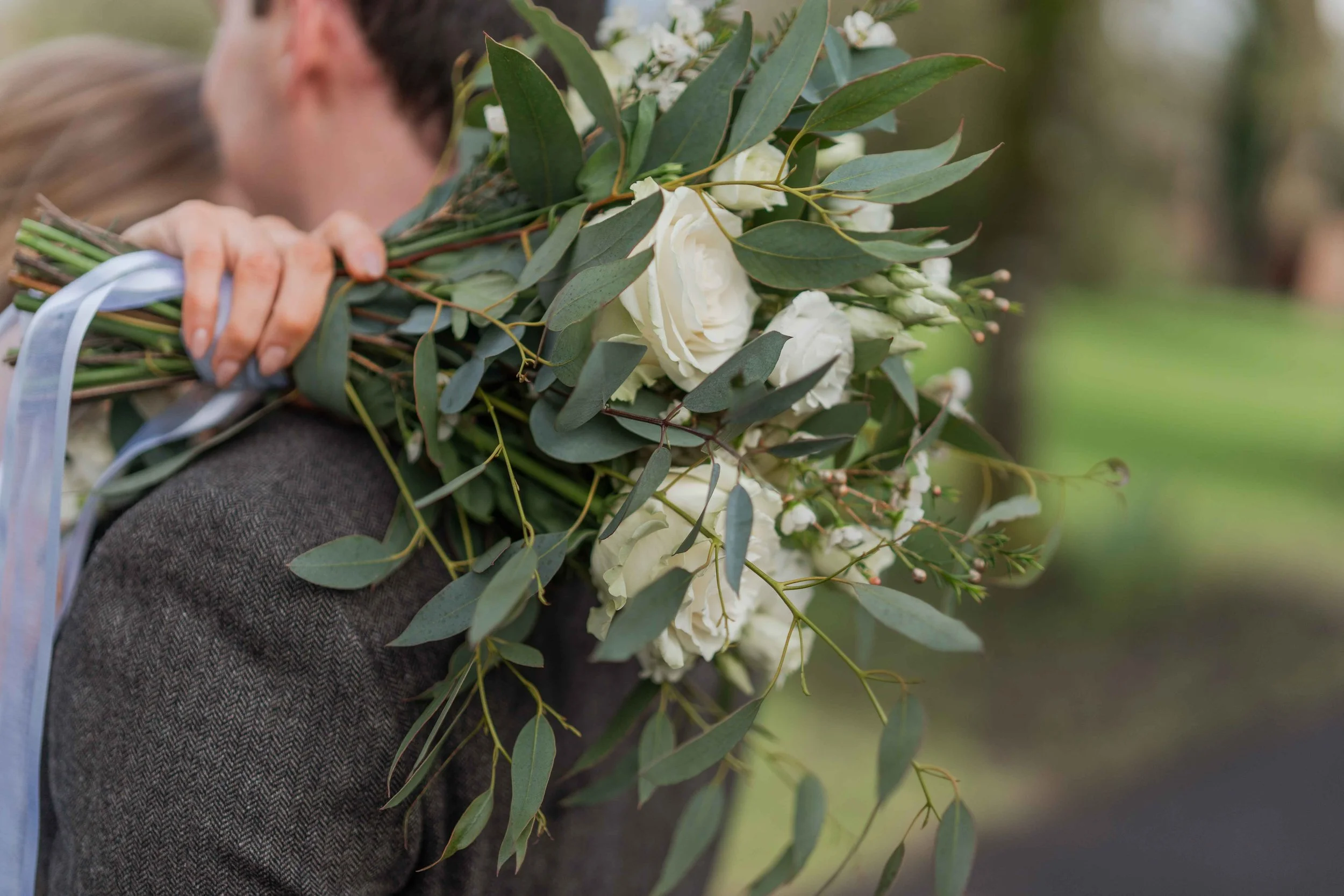 Person holding a large bouquet of white roses, greenery, and small white flowers on their shoulder