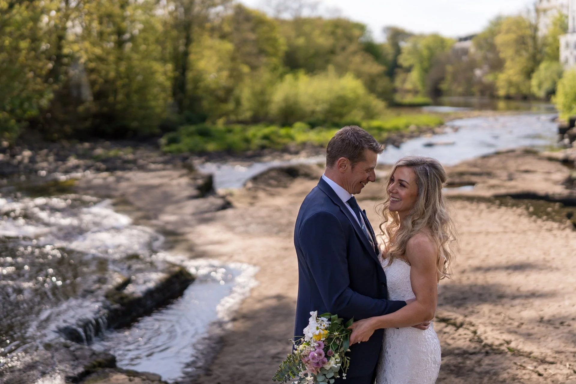A newlywed couple stands close together near a river on a sunny day, with trees and buildings in the background.