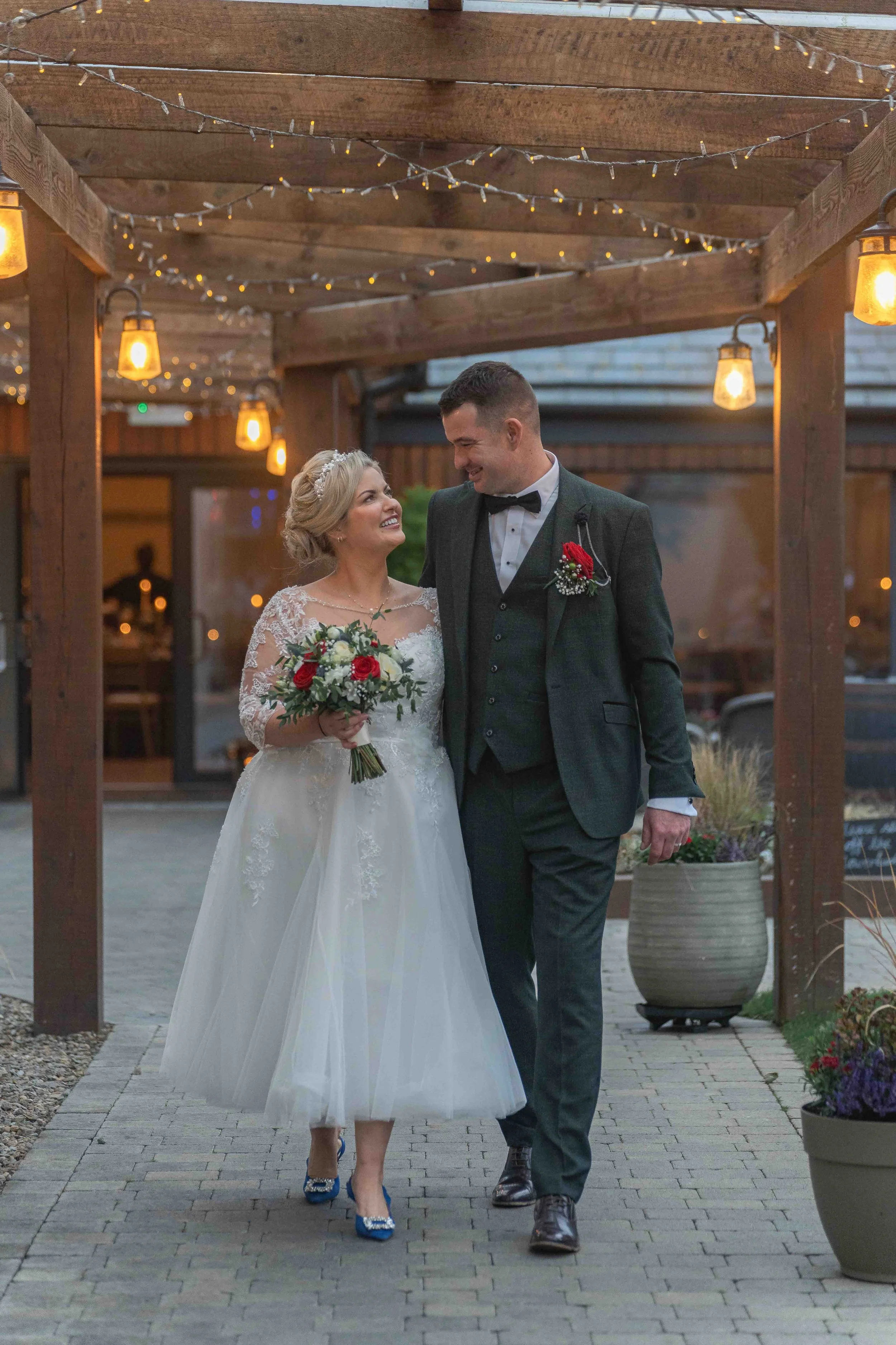 A bride and groom walking together outdoors, smiling at each other, under a wooden pergola decorated with string lights. The bride is in a white wedding dress holding a bouquet, and the groom is in a dark suit with a bow tie.
