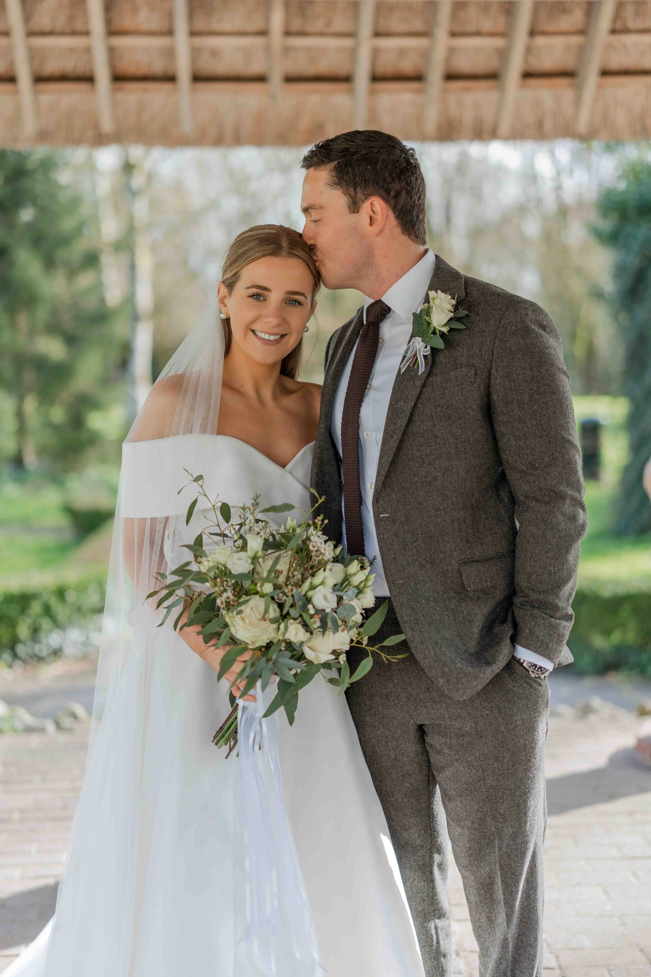 A newlywed couple on their wedding day, with the groom kissing the bride on the forehead.