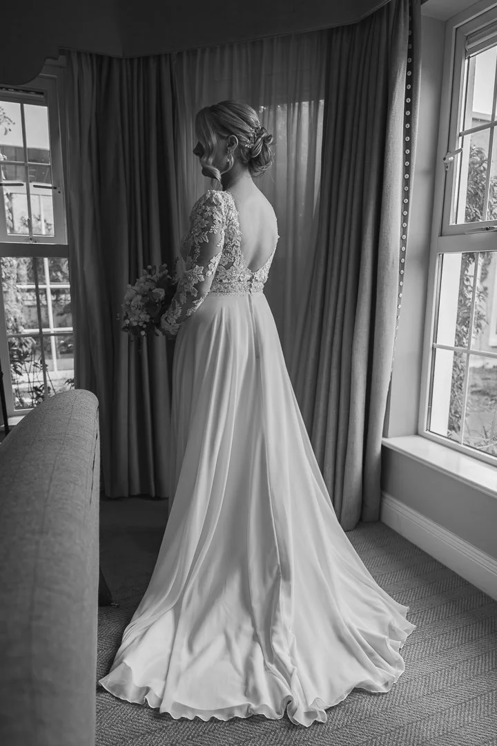 A bride in a wedding dress holding a bouquet, standing near a window indoors, captured in black and white.