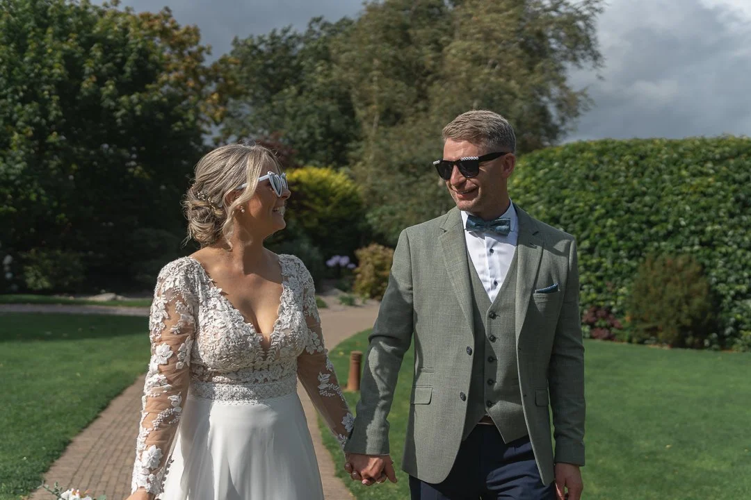 Bride and groom holding hands and smiling at each other outdoors on a sunny day.