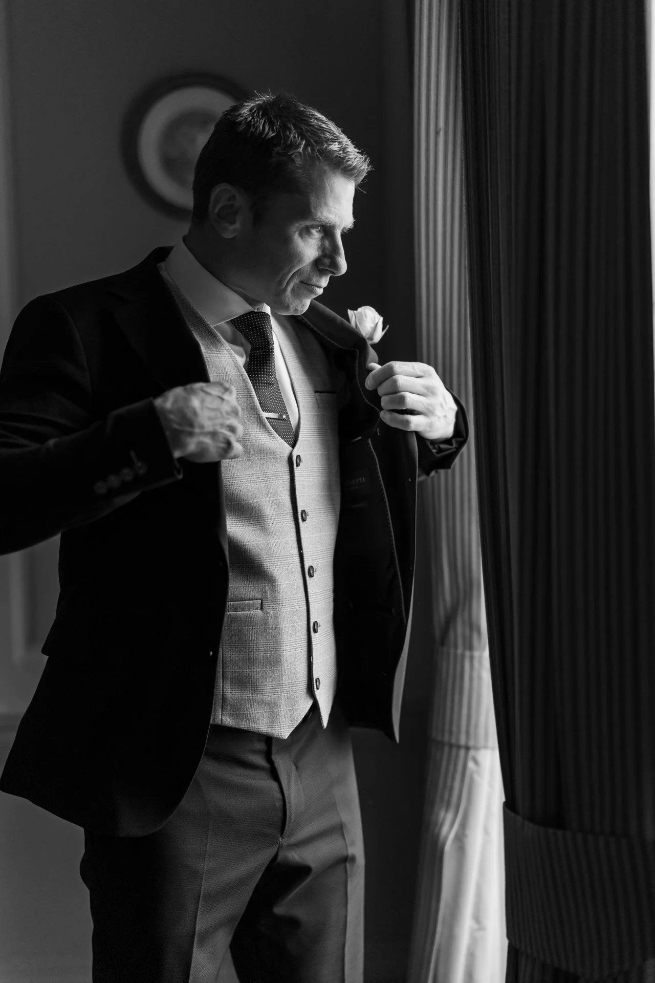 A man in formal attire, adjusting his jacket and looking out a window, in a black-and-white photo.