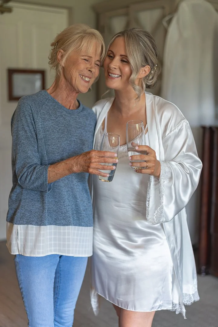 An elderly woman and a middle-aged woman, both smiling, sharing a toast with glasses of water in a cozy room.
