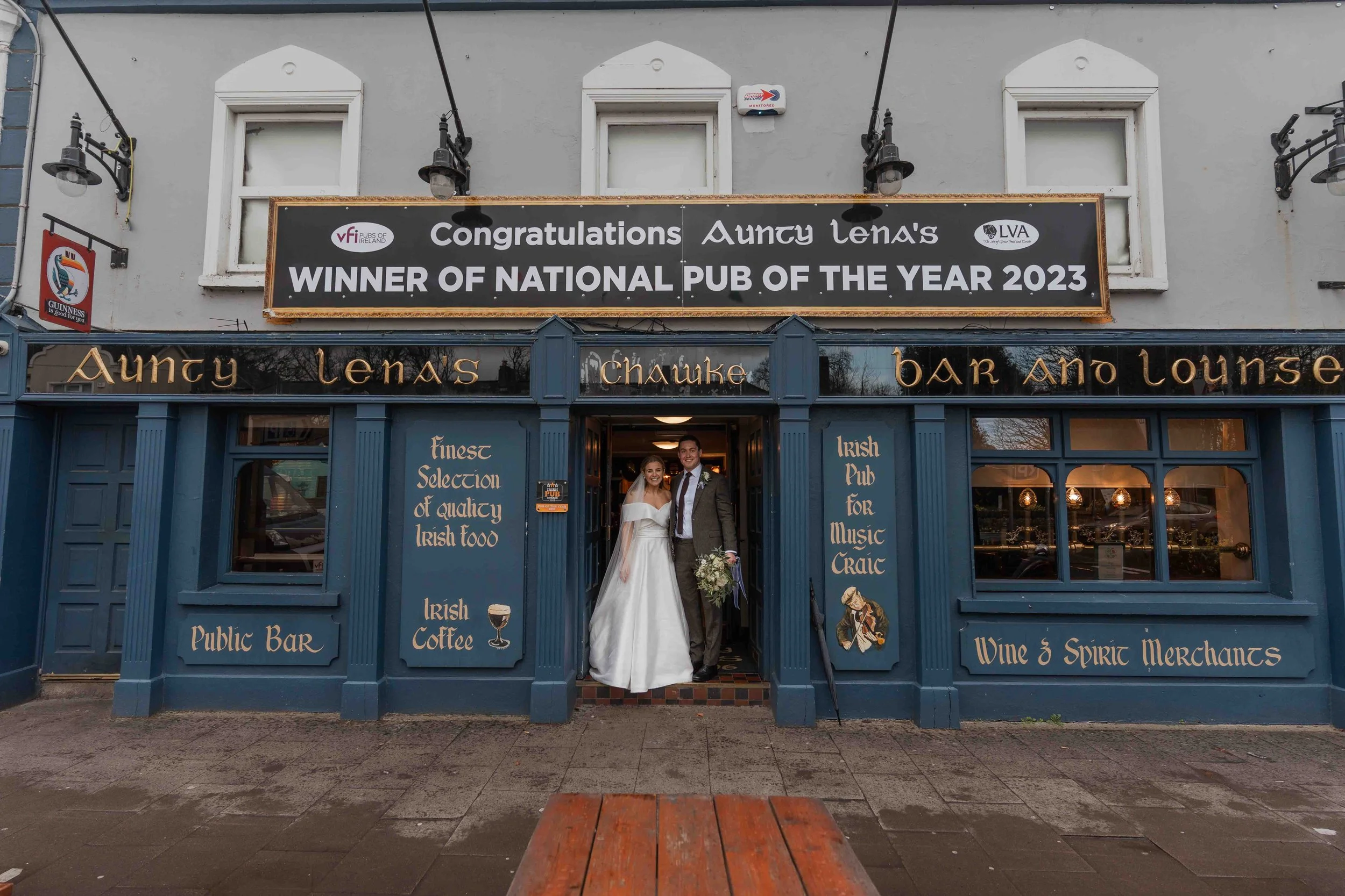 A wedding couple standing at the entrance of a pub and lounge called Aunty Lena's, celebrating winning the 2023 National Pub of the Year. The bride and groom are smiling, with the bride wearing a white dress and the groom in a suit holding a bouquet 