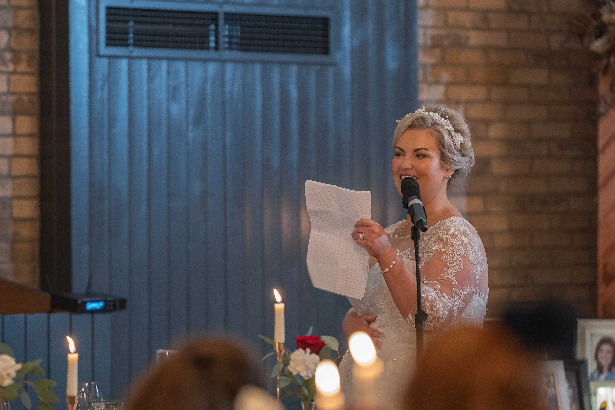 Bride giving a speech at her wedding reception, holding a piece of paper and standing in front of a microphone, with lit candles and floral arrangements on the table in the foreground.