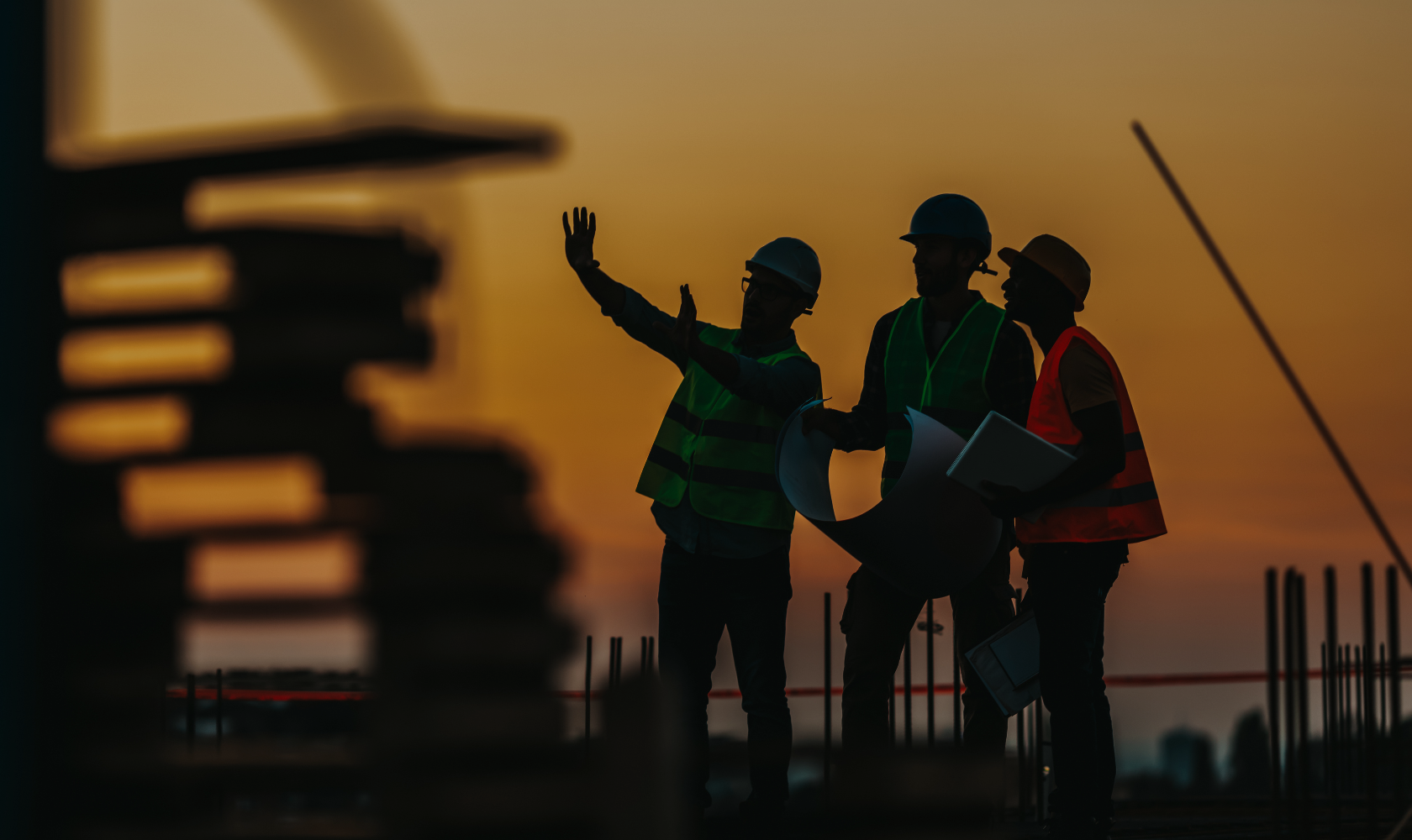 Silhouettes of three construction workers on a building site at sunset, wearing hard hats and reflective vests, with one gesturing and holding plans, in a city skyline environment.
