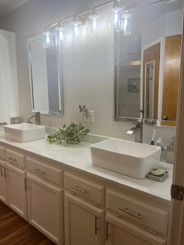Bathroom vanity with dual rectangular vessel sinks, white cabinetry, a marble countertop, a plant, and a large mirror with lights above.