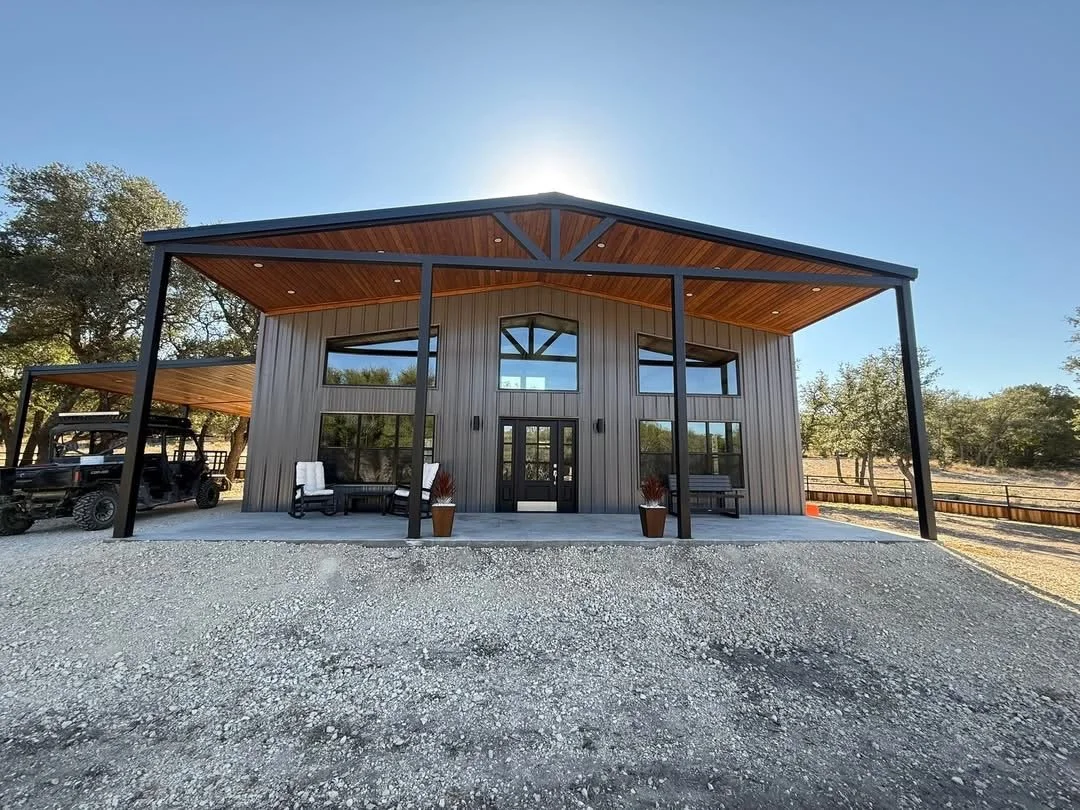 Modern house with black metal framing, vertical siding, and large windows, set on a gravel foundation with exterior seating, surrounded by trees.