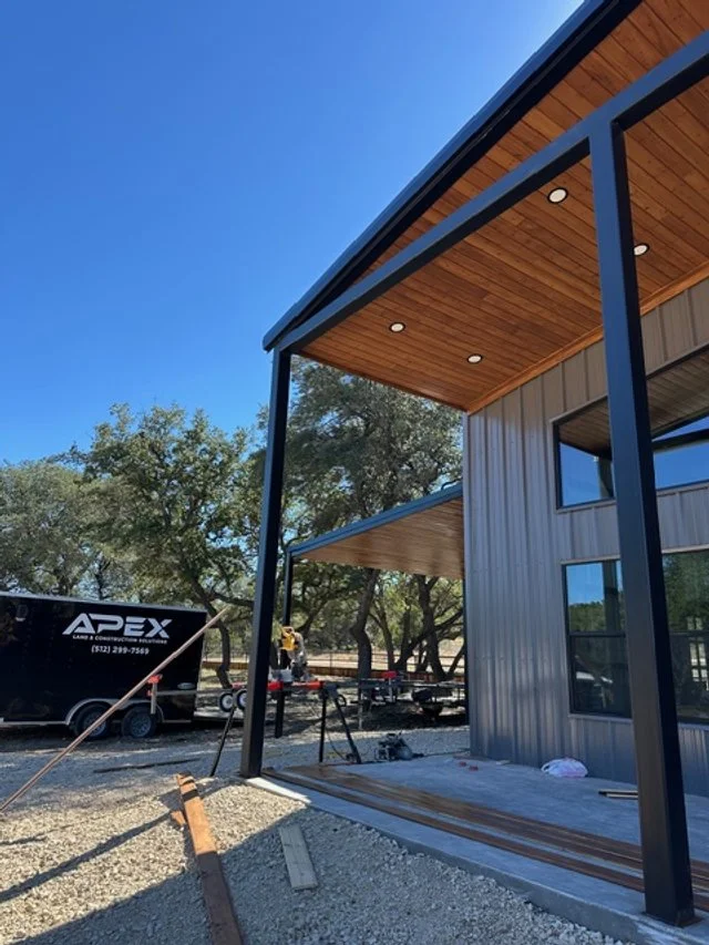 Construction site with a modern metal and wood building, black metal framing, and a covered porch area, surrounded by trees and construction equipment.