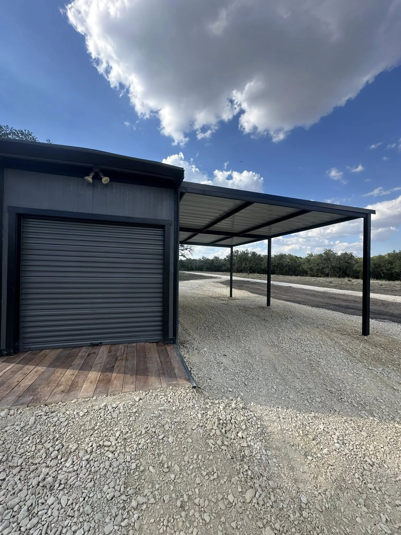 A black building with a garage door and a covered carport on a gravel driveway, with trees and a cloudy sky in the background.