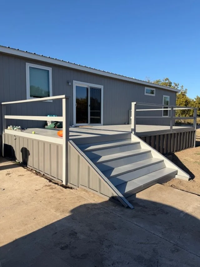 A gray manufactured home with a newly built gray wooden deck and stairs leading up to the sliding glass door. The deck has a railing, and the ground around it is bare dirt. The sky is clear and blue.
