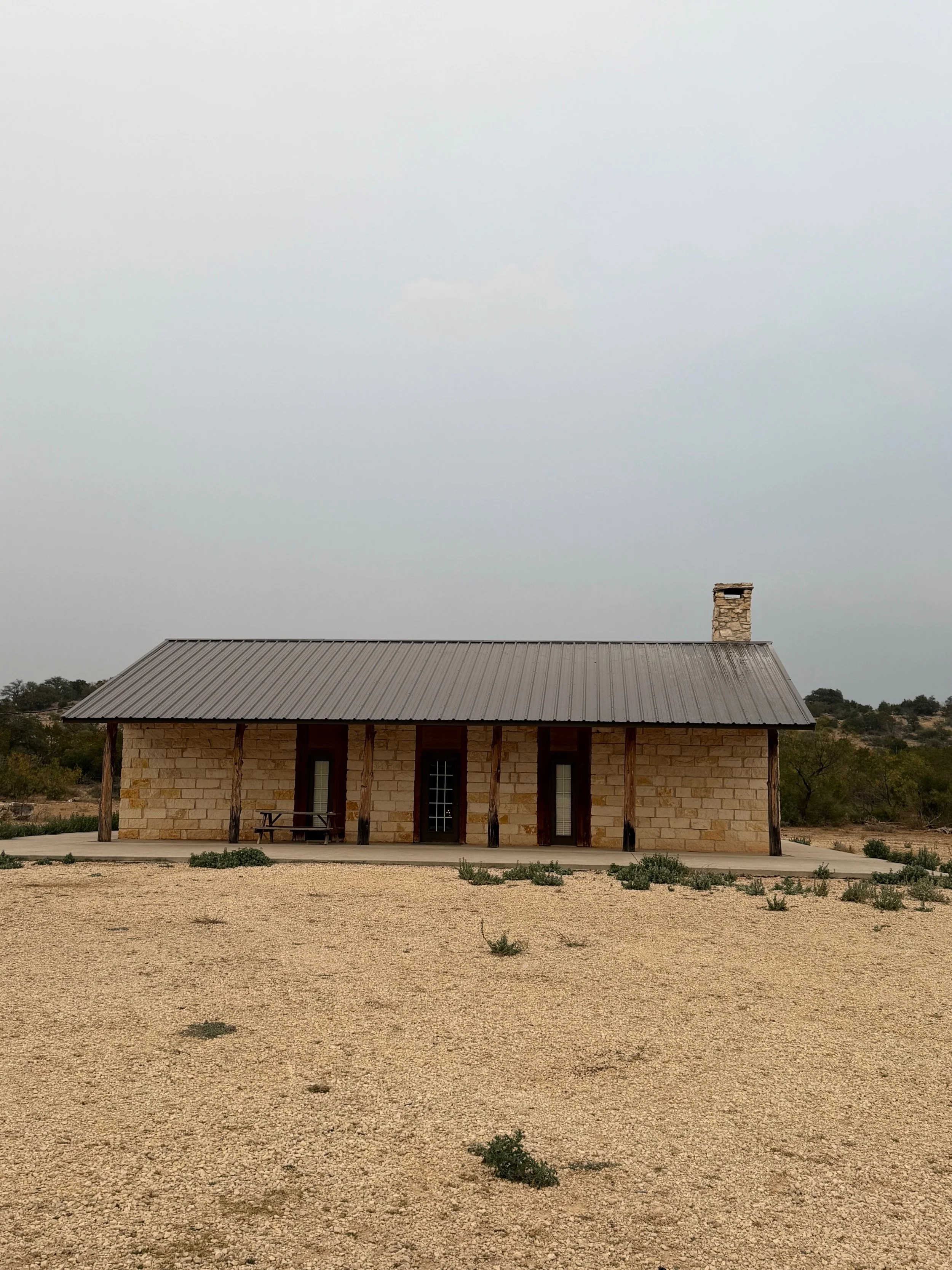 A small, rustic house in a desert landscape with a metal roof, stone walls, wooden pillars, and a chimney, surrounded by sparse vegetation and an open sandy area.