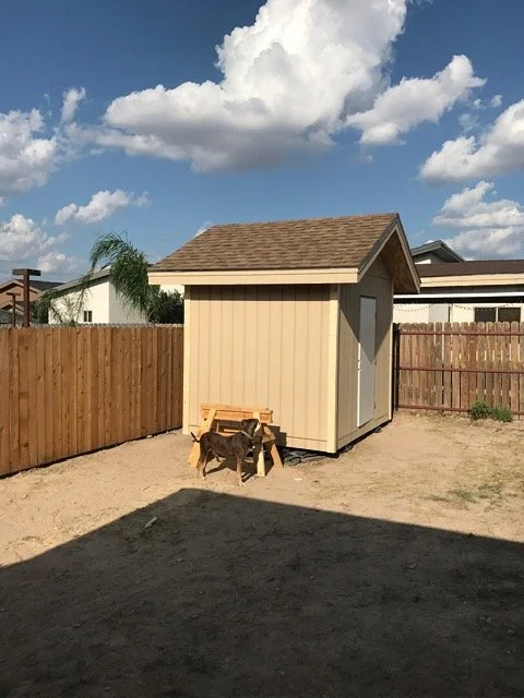 Backyard with a small beige shed, a wooden bench, and a dog in front, enclosed by a wooden fence, under a partly cloudy sky.