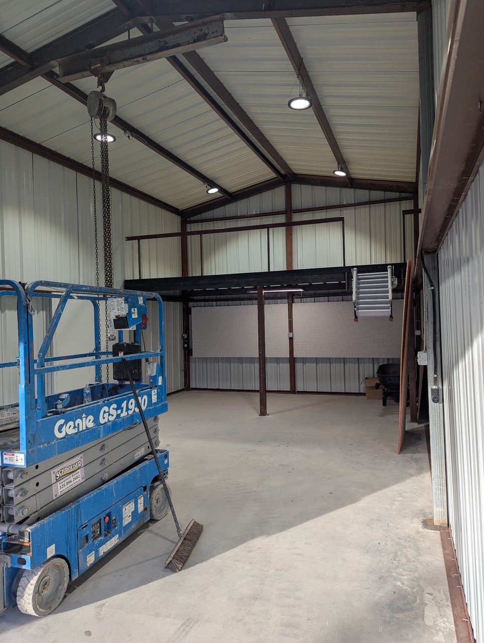 Empty industrial warehouse with a blue Genie scissor lift and a broom on the concrete floor, metal walls, and a lofted platform with a ladder.