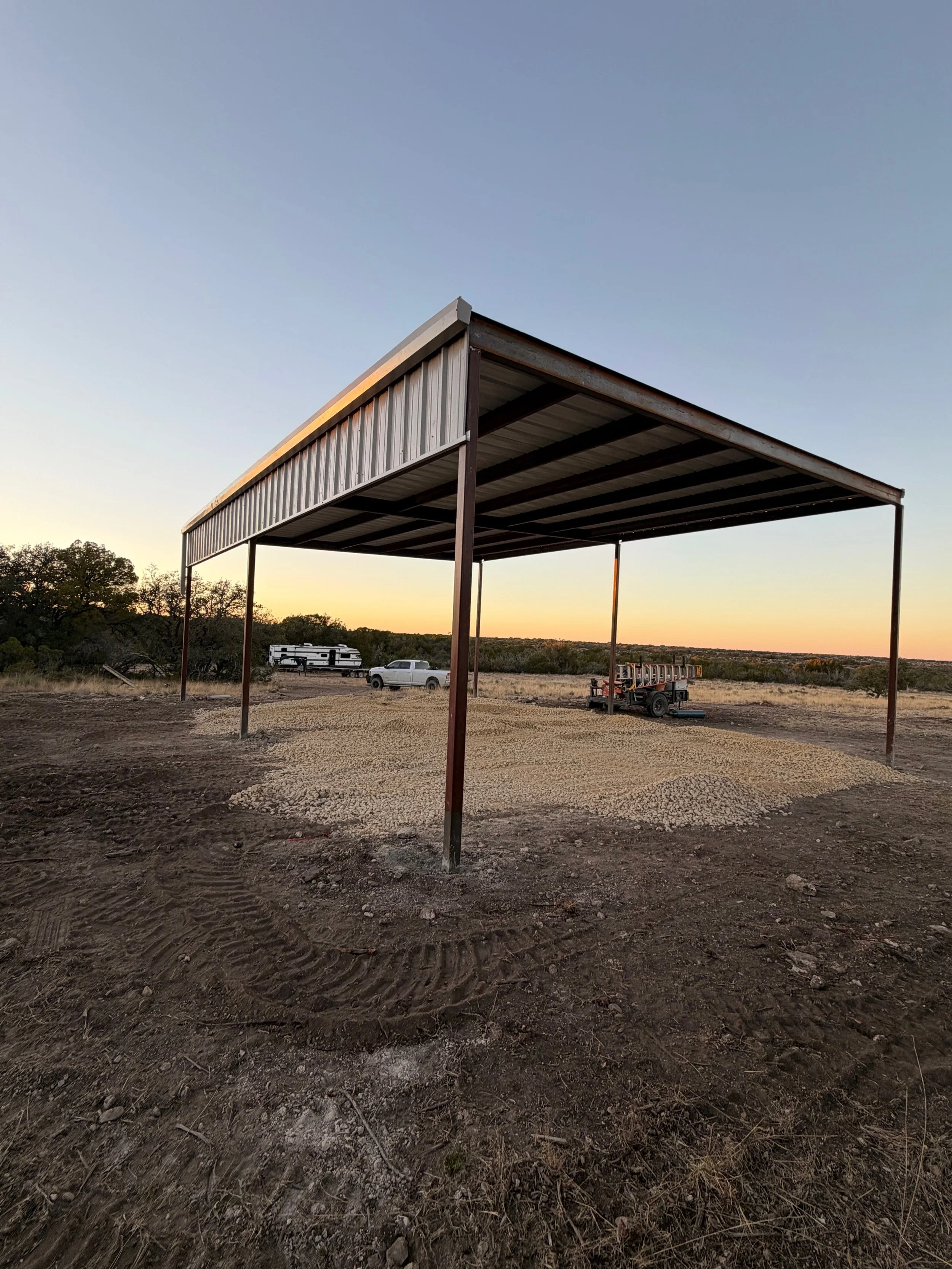 A metal shelter in a rural area with a gravel floor, surrounded by dirt and sparse vegetation, with a pickup truck, a trailer, and a camper in the background at sunset.