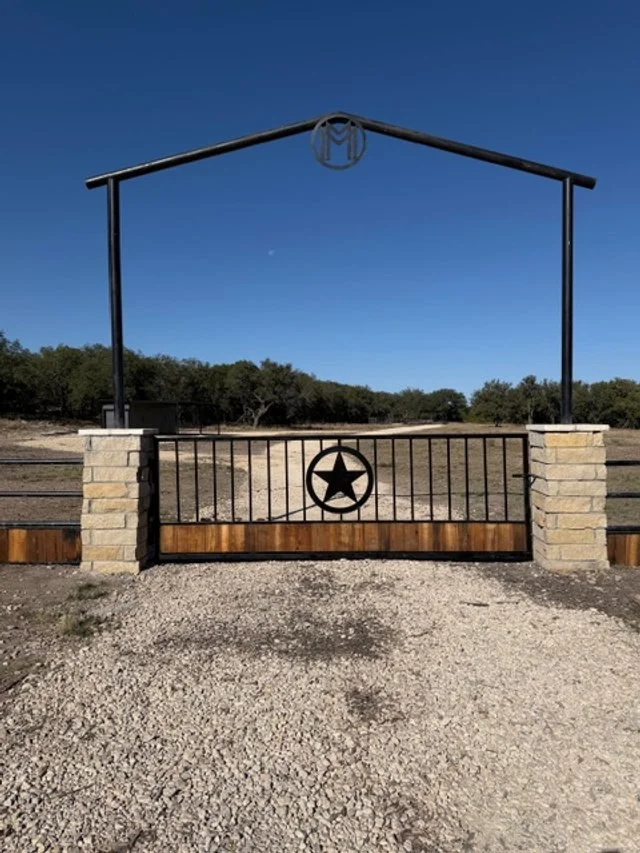 A black metal gate with a star in a circle at the center, supported by stone pillars and topped with a metal arch, leading to a dirt pathway in a rural area with trees in the background under a clear blue sky.