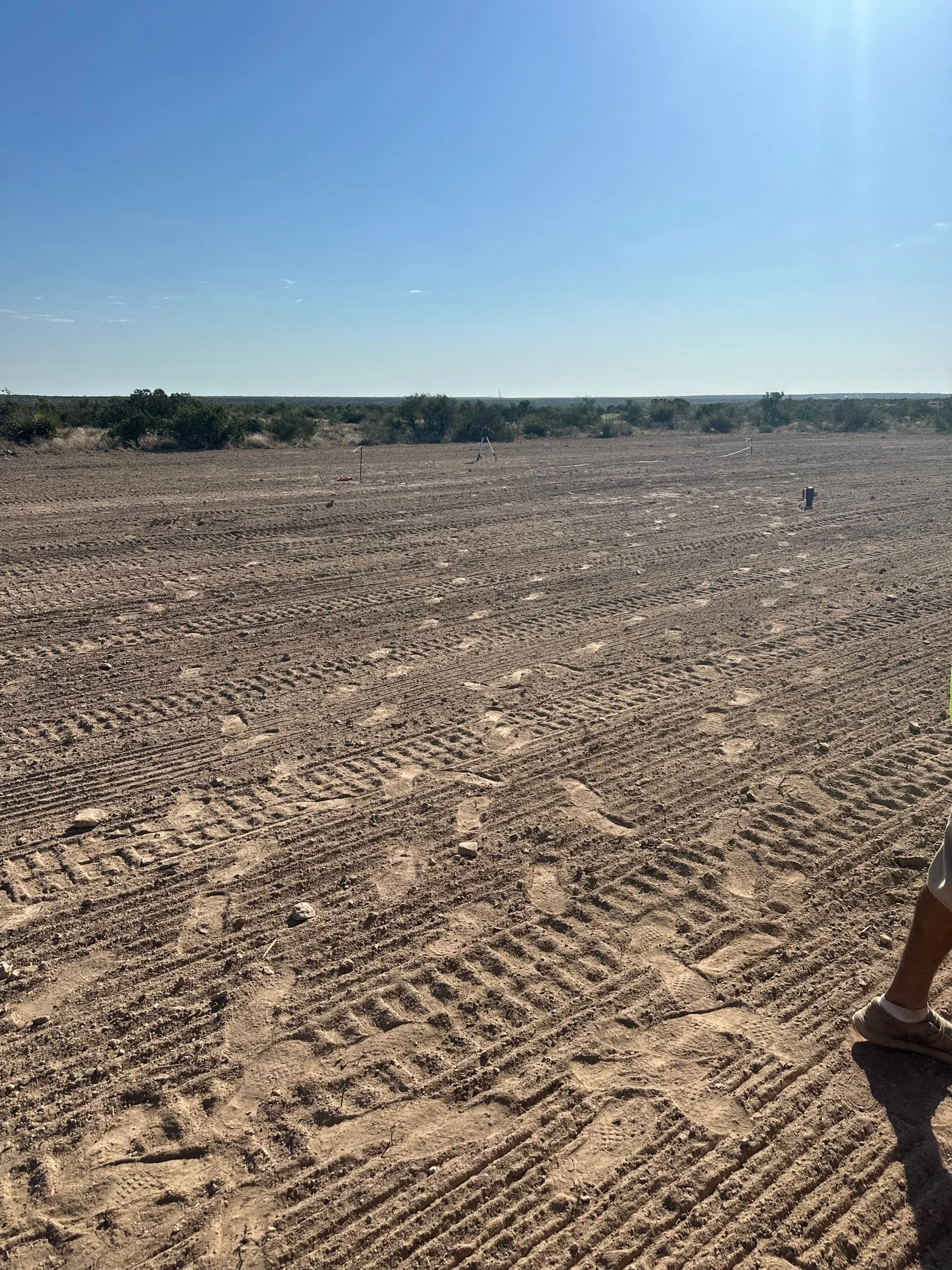 A wide view of a desert landscape with cleared, flat sandy ground, sparse bushes in the distance, and a clear blue sky. Part of a person's leg and shoe is visible on the lower right corner.