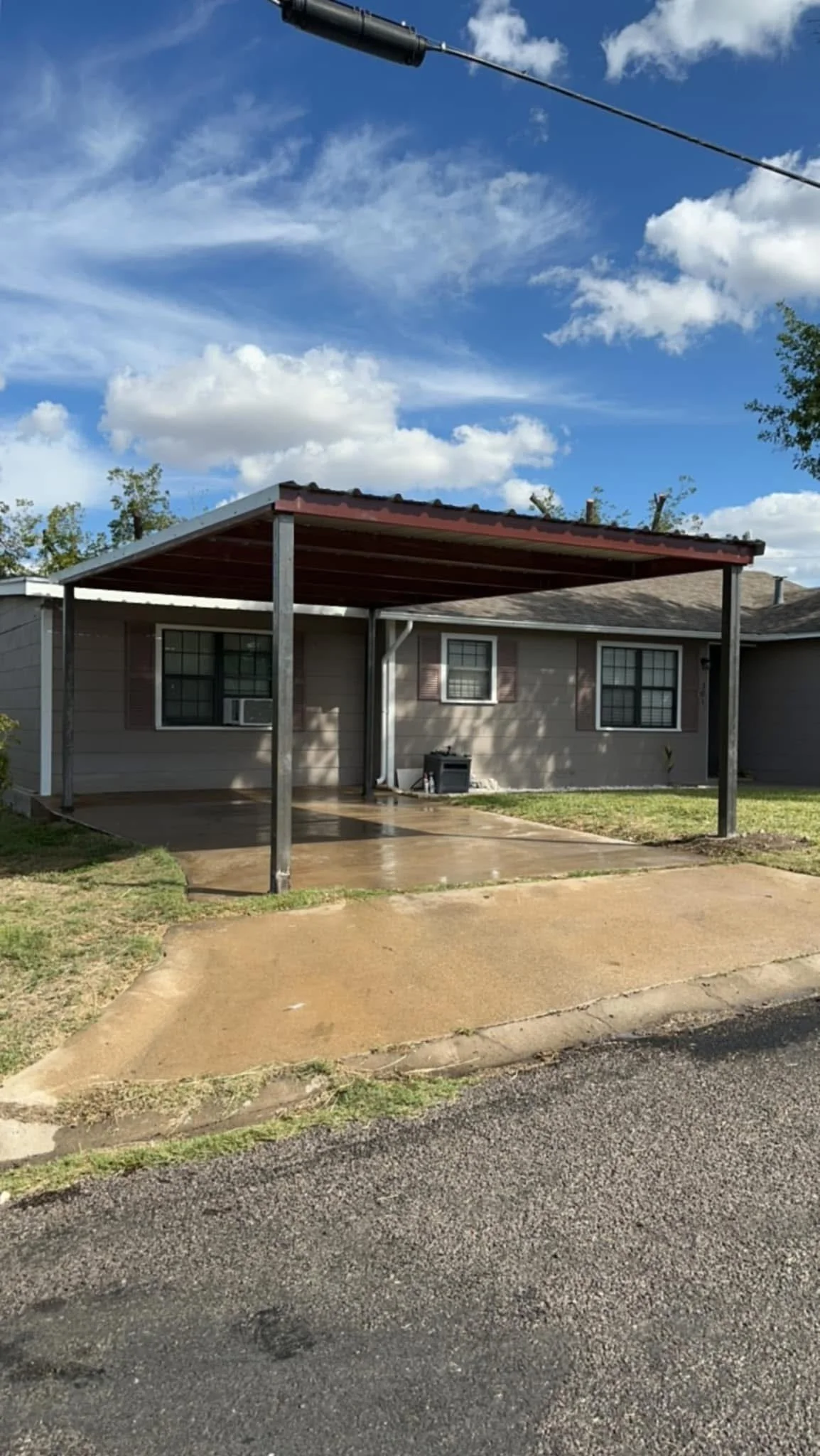 A house with a covered patio area, concrete floor, and a grassy yard, under a blue sky with clouds.