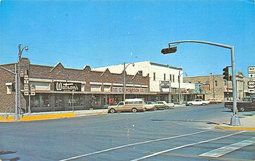 Street view of a small town intersection with vintage cars and storefronts, including Watson's, C. Morrison Co., a bakery, and a drugstore, under a clear blue sky.