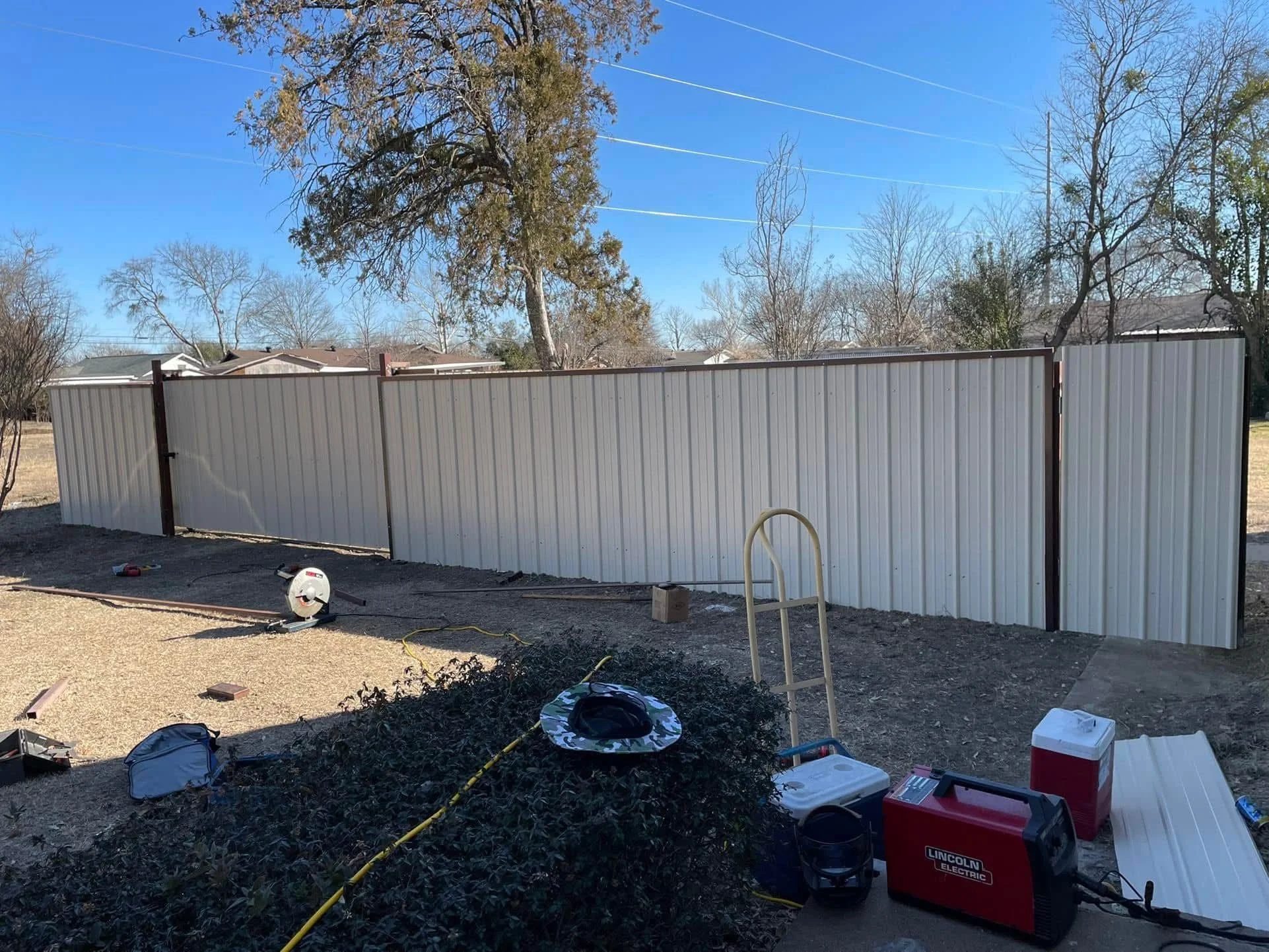 A backyard with a white metal fence, construction tools, and equipment on the ground, with leafless trees and houses in the background under a clear blue sky.