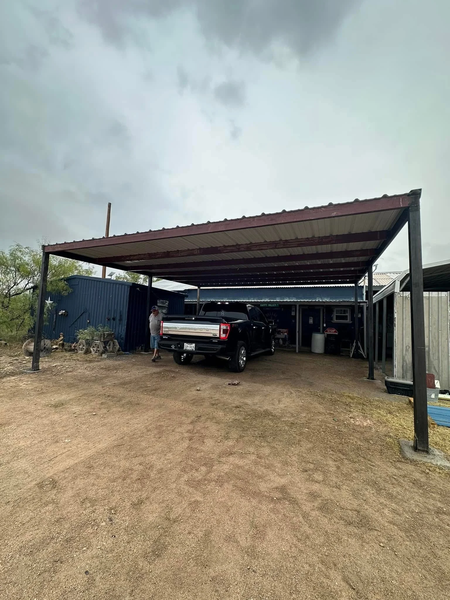 A covered carport with a black pickup truck and a man standing nearby, in a rural setting with dirt ground and some outdoor objects.