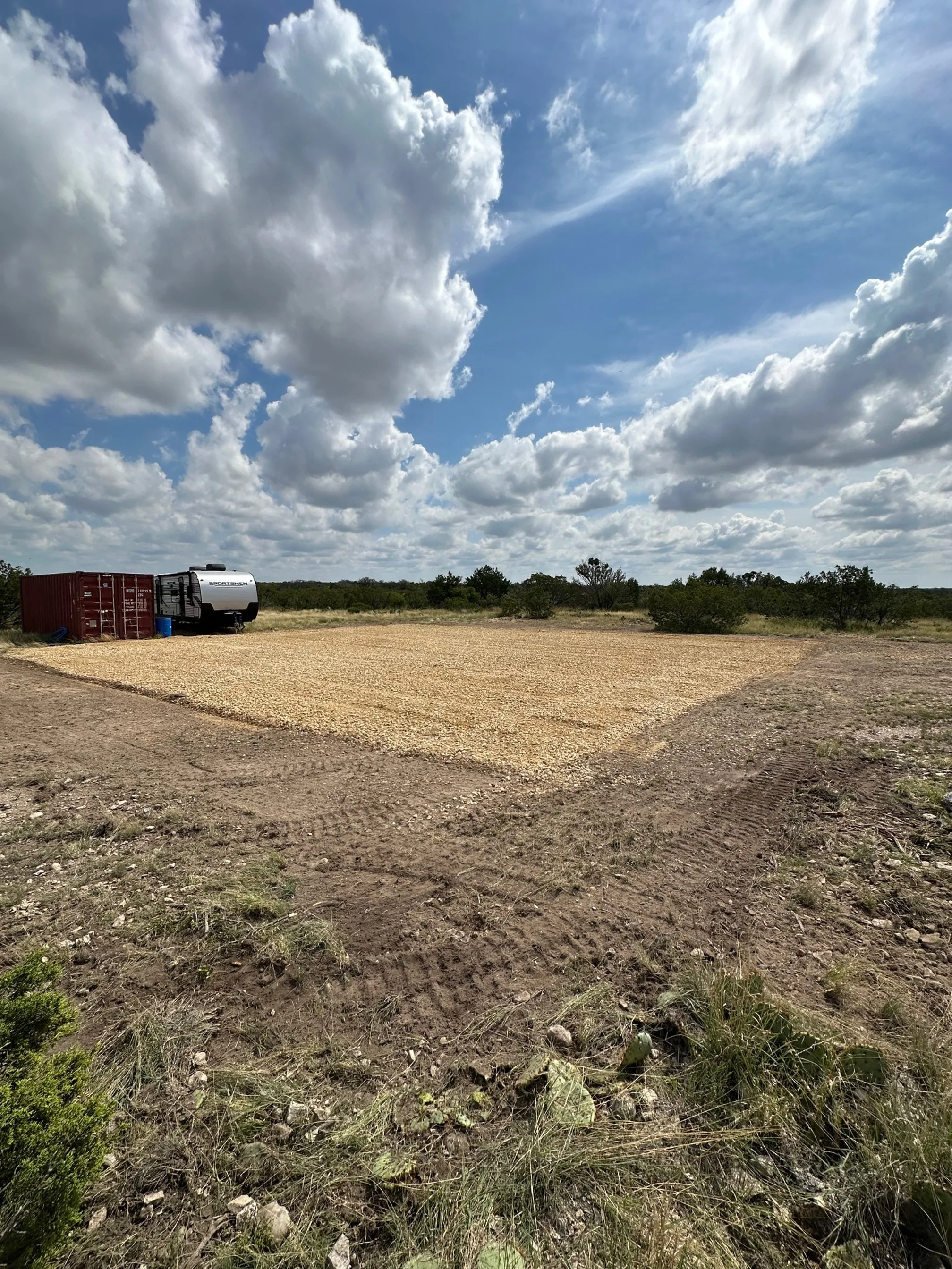 A gravelly outdoor area with a cleared patch of land, tire tracks in the dirt, a storage container, and a small RV trailer under partly cloudy sky with trees in the background.