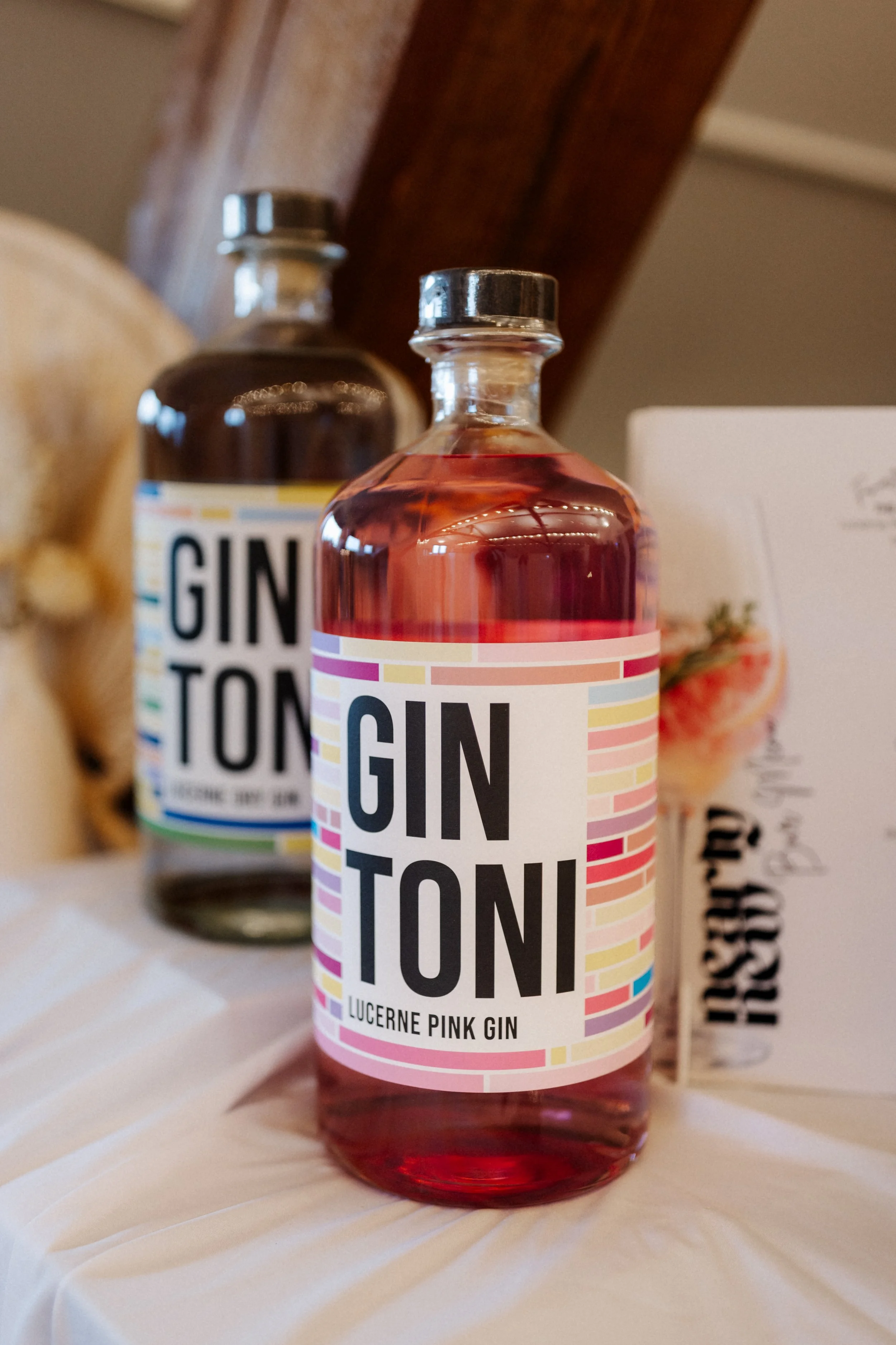 Eine Flasche mit pinkfarbenem Gin mit dem Label 'Lucerne Pink Gin' und die Markenbezeichnung 'Gintonic'. Im Hintergrund sind weitere Flaschen und Verpackungen sichtbar.