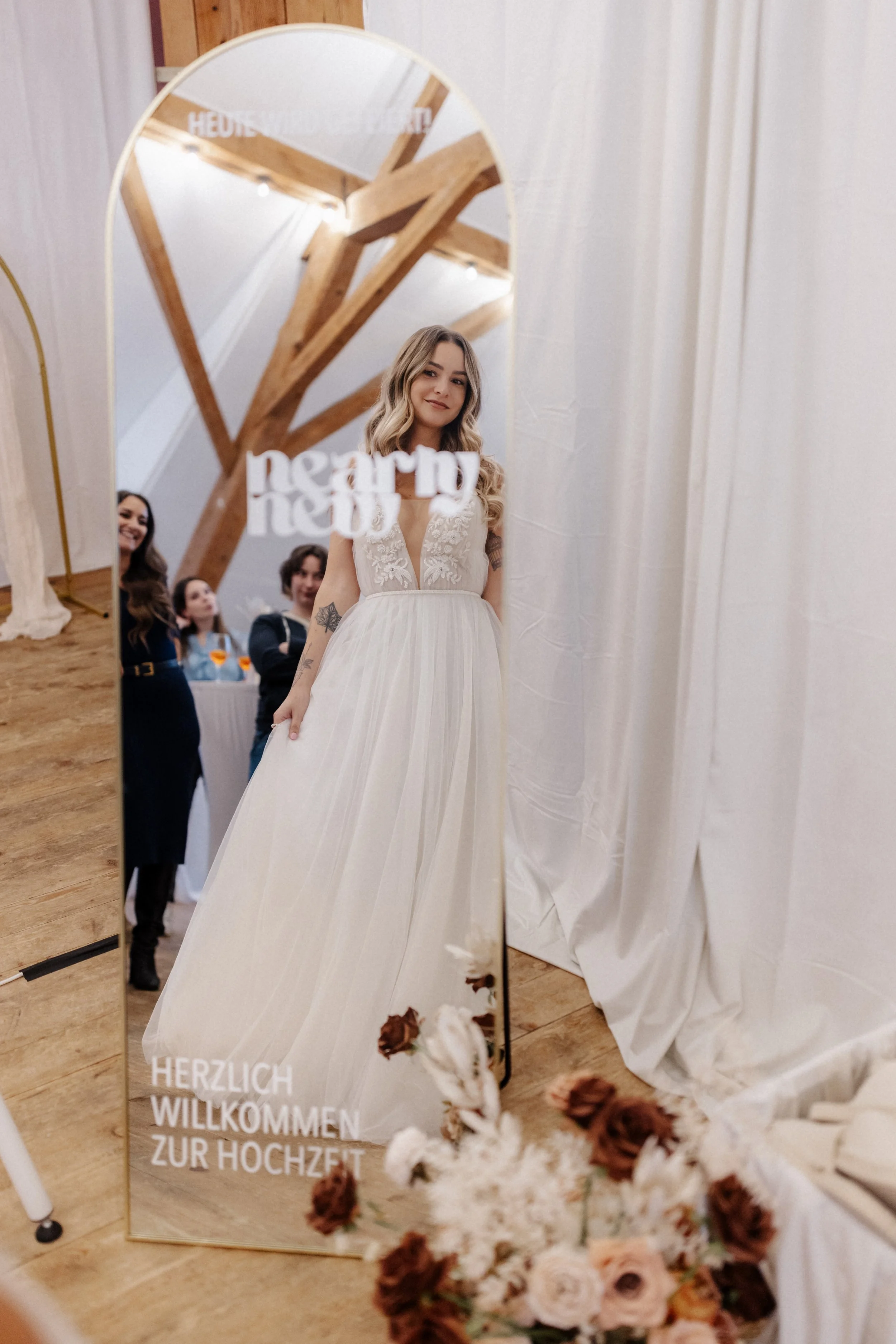 Braut in weißem Brautkleid wird in einem Raum vor einem Spiegel fotografiert. Im Hintergrund sind zwei Frauen und ein Mann. Der Spiegel hat einen goldenen Rahmen und trägt die Beschriftung: 'Herzlich willkommen zur Hochzeit'. Im Vordergrund sind Blumen im Weiß- und Rosaton sichtbar.