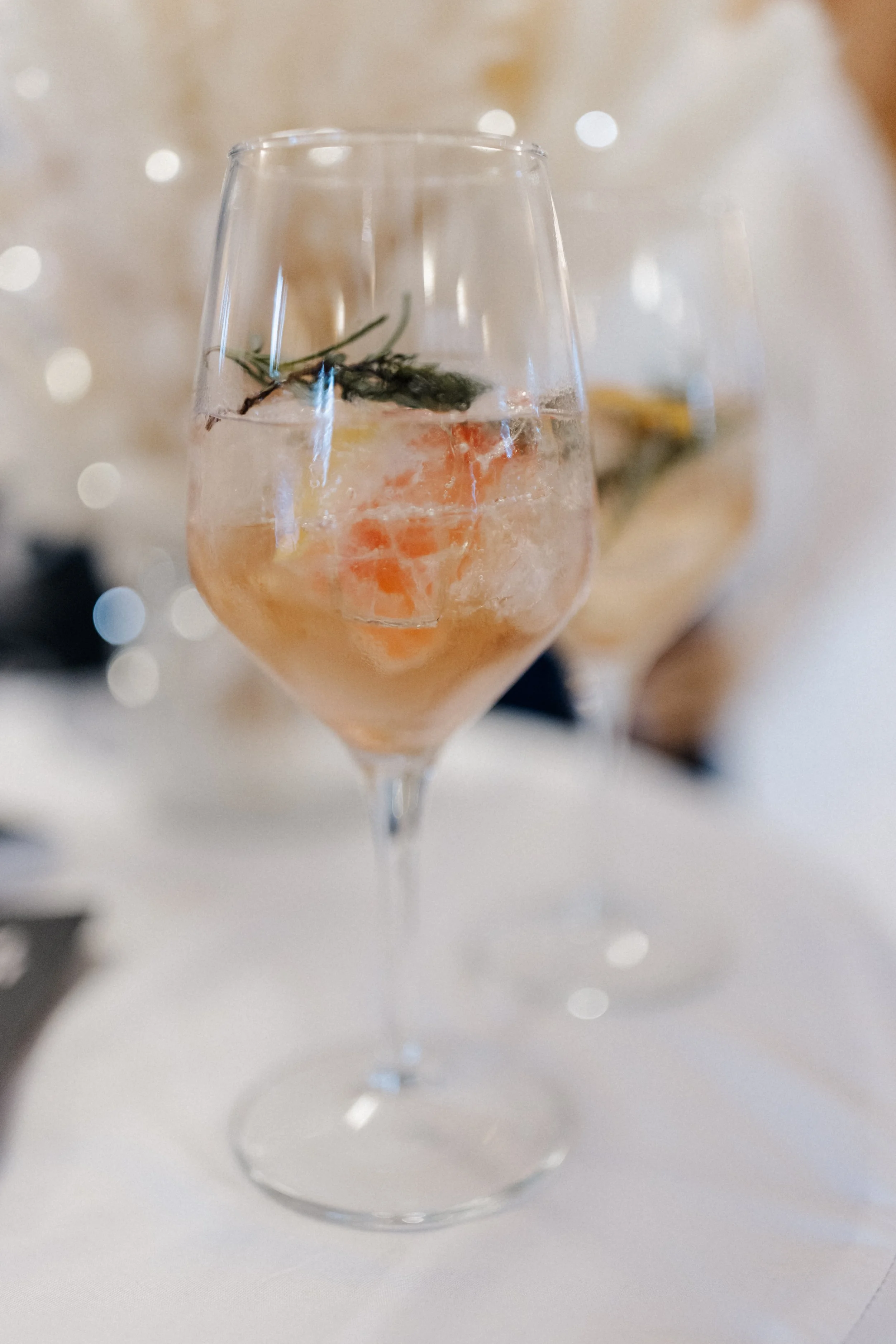 Ein Weinglas mit Wasser, einem Zweig Rosmarin und einem Stück Grapefruit darin, auf einem weißen Tisch, unscharfer Hintergrund.