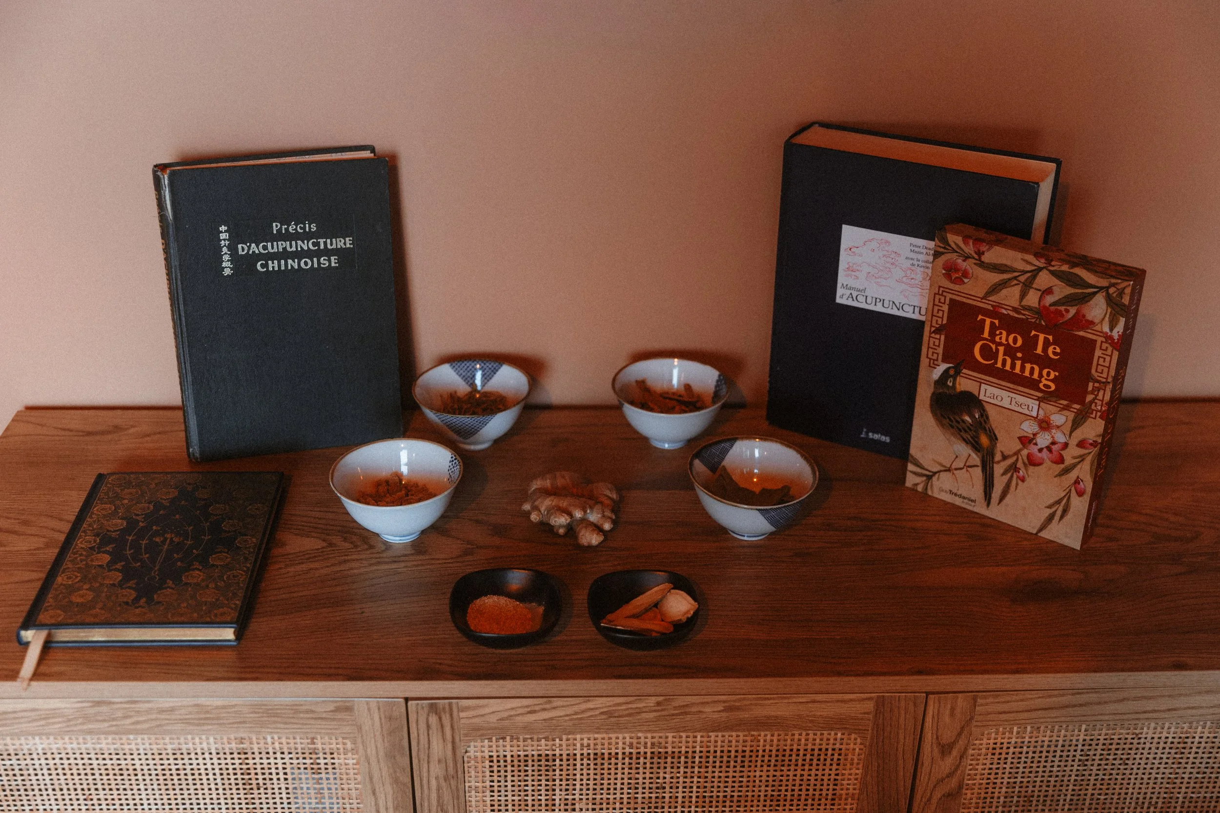 Une table en bois avec plusieurs livres sur l'acupuncture chinoise, quatre petits bols blancs contenant des herbes, deux petits bols noirs avec des épices, un livre de Tamarind et deux livres plus grands sur l'acupuncture et la philosophie chinoise.
