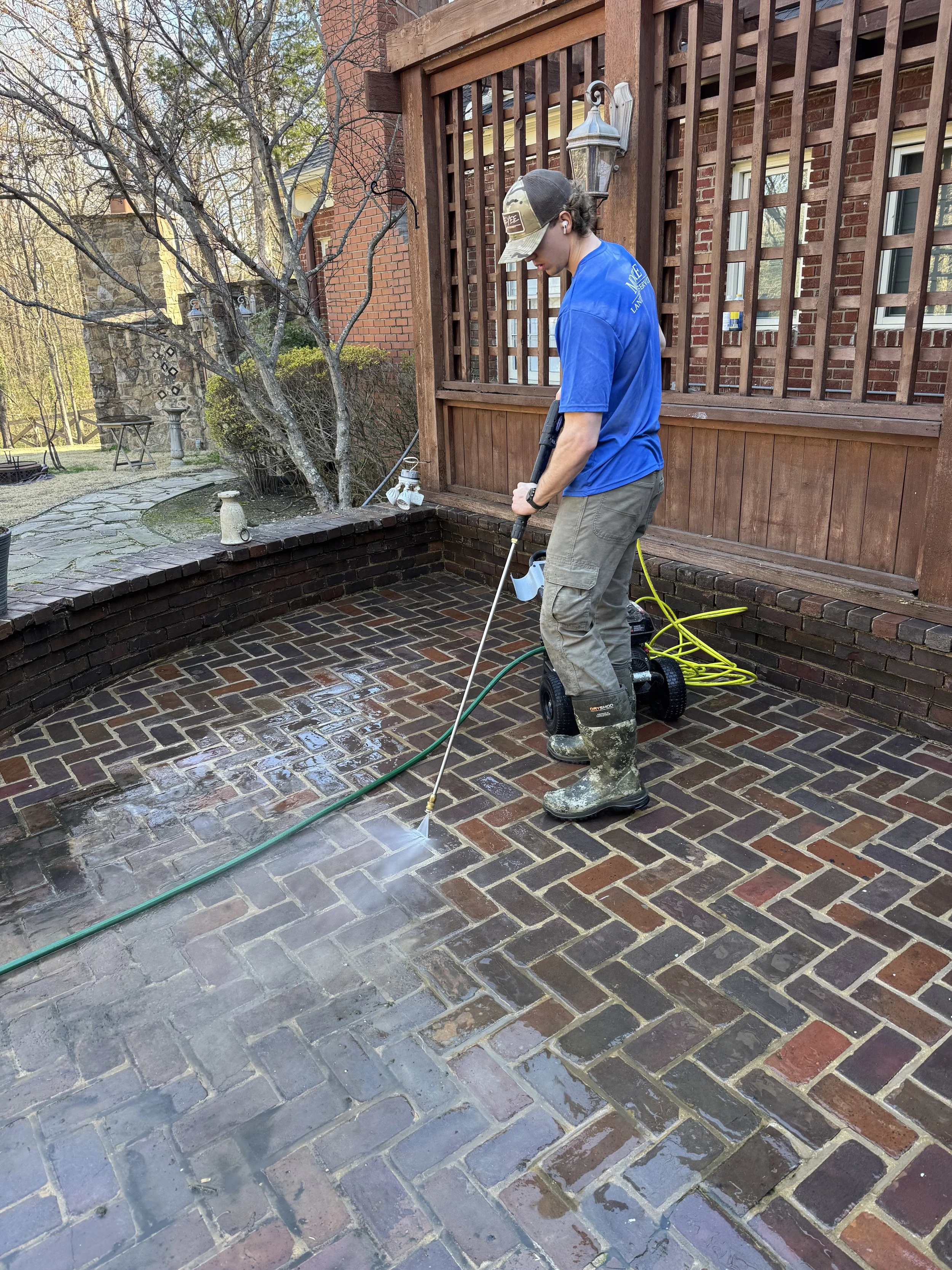 A person using a power washer to clean a brick patio outside a house with a wooden porch.
