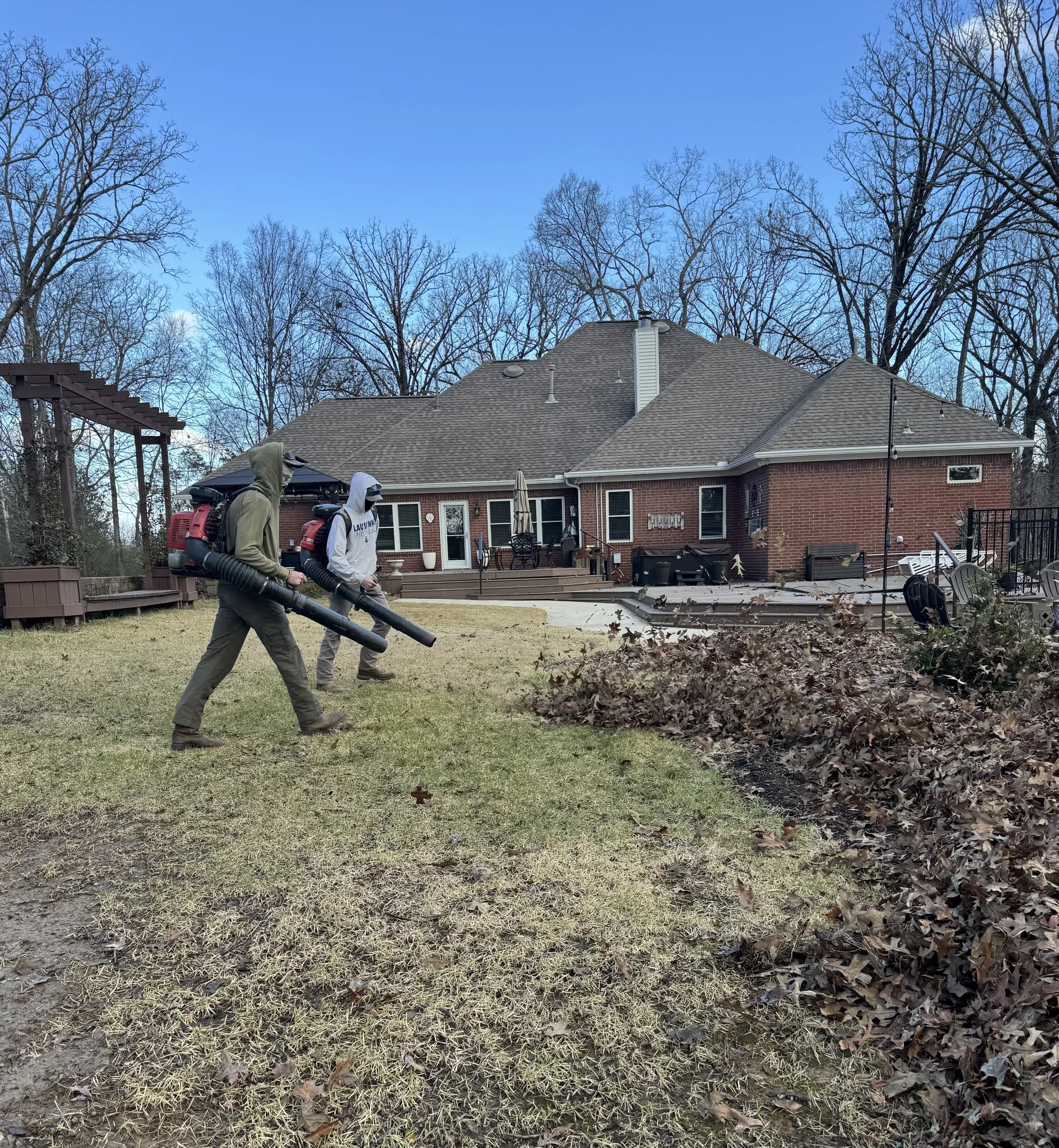 Two people wearing backpacks and hooded jackets, using leaf blowers to clear fallen leaves from a backyard lawn, with a brick house and leafless trees in the background.