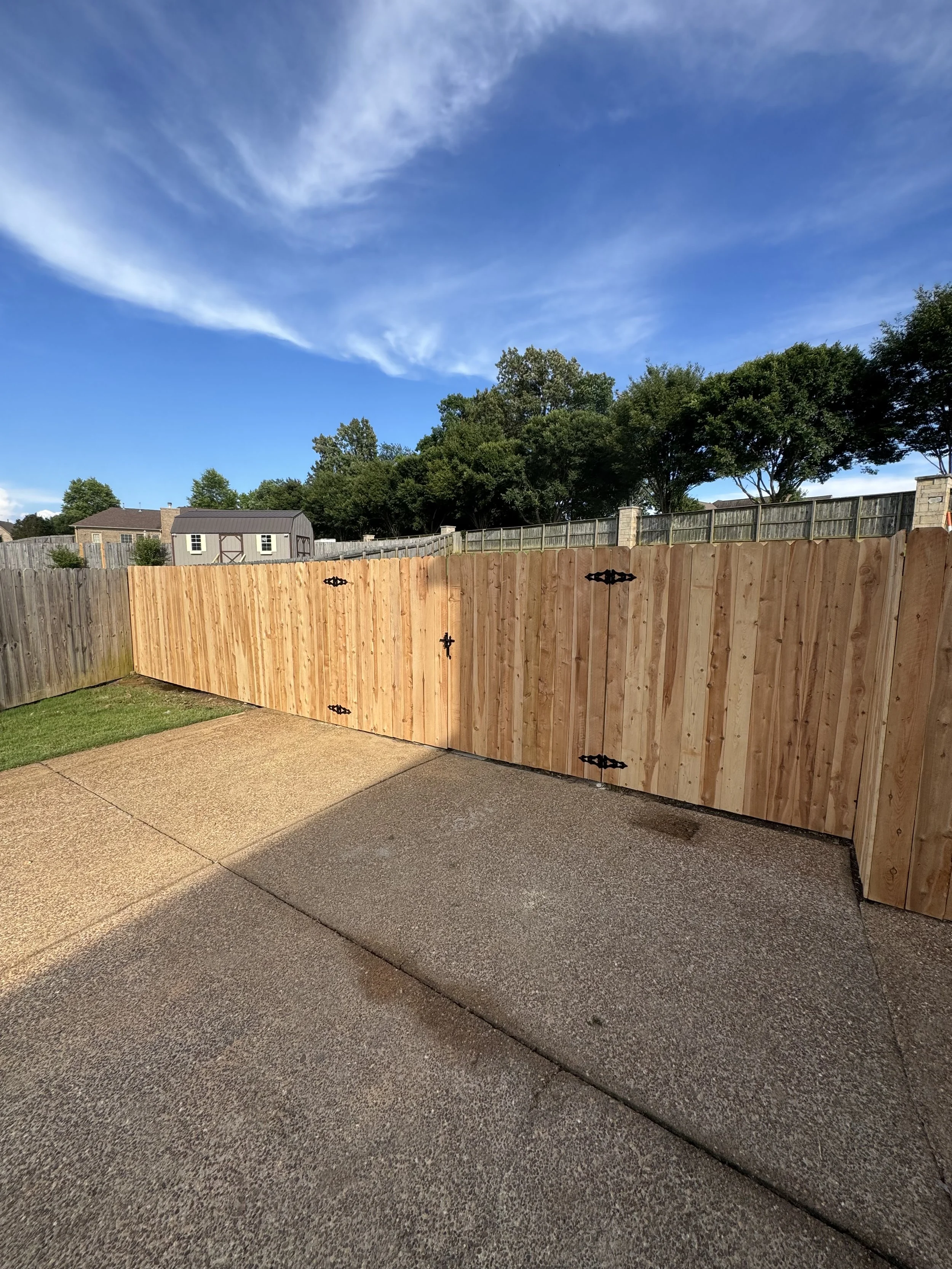 A backyard with a new wooden fence, concrete patio, some grass, and a blue sky with clouds.