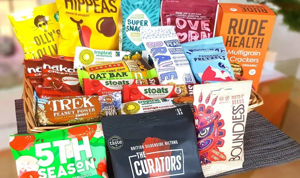 Basket filled with various snack packs and boxes, including chips, crackers, energy bars, and gummies. Some of the brands visible are Olly's Olives, Hippeas, Love Corn, Rude Health, Nakd, Tropeaka, The Curators, and Underlings.