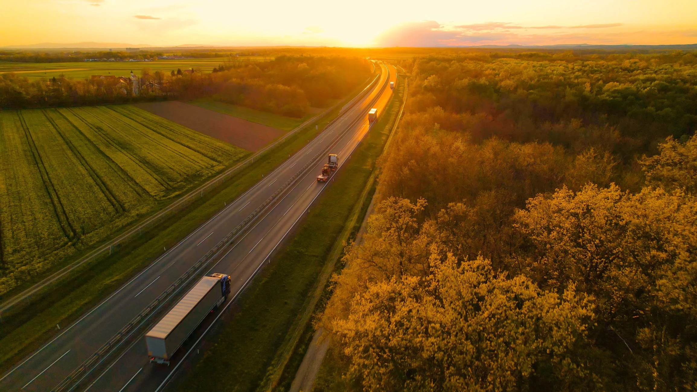 Aerial view of a highway at sunset, with trucks traveling between green fields and forests that glow in warm light.