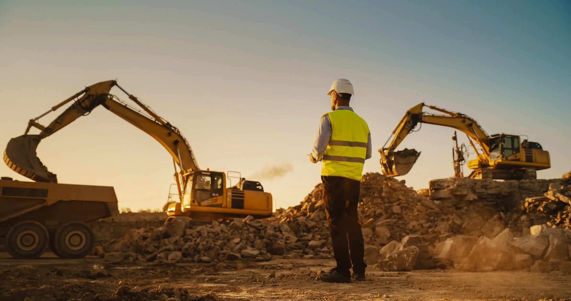 A construction worker in a helmet and reflective vest observes two excavators moving rubble at sunset.