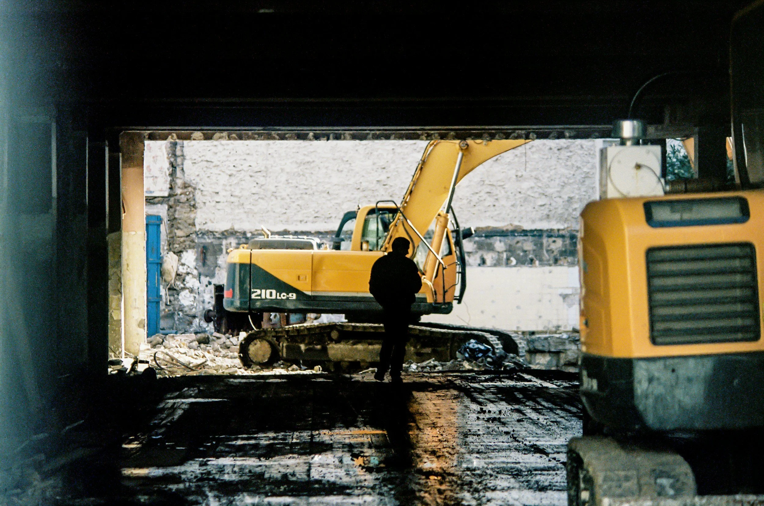 Silhouette of a person standing in a dimly lit construction site, with two large yellow excavators and a partially demolished wall in view.