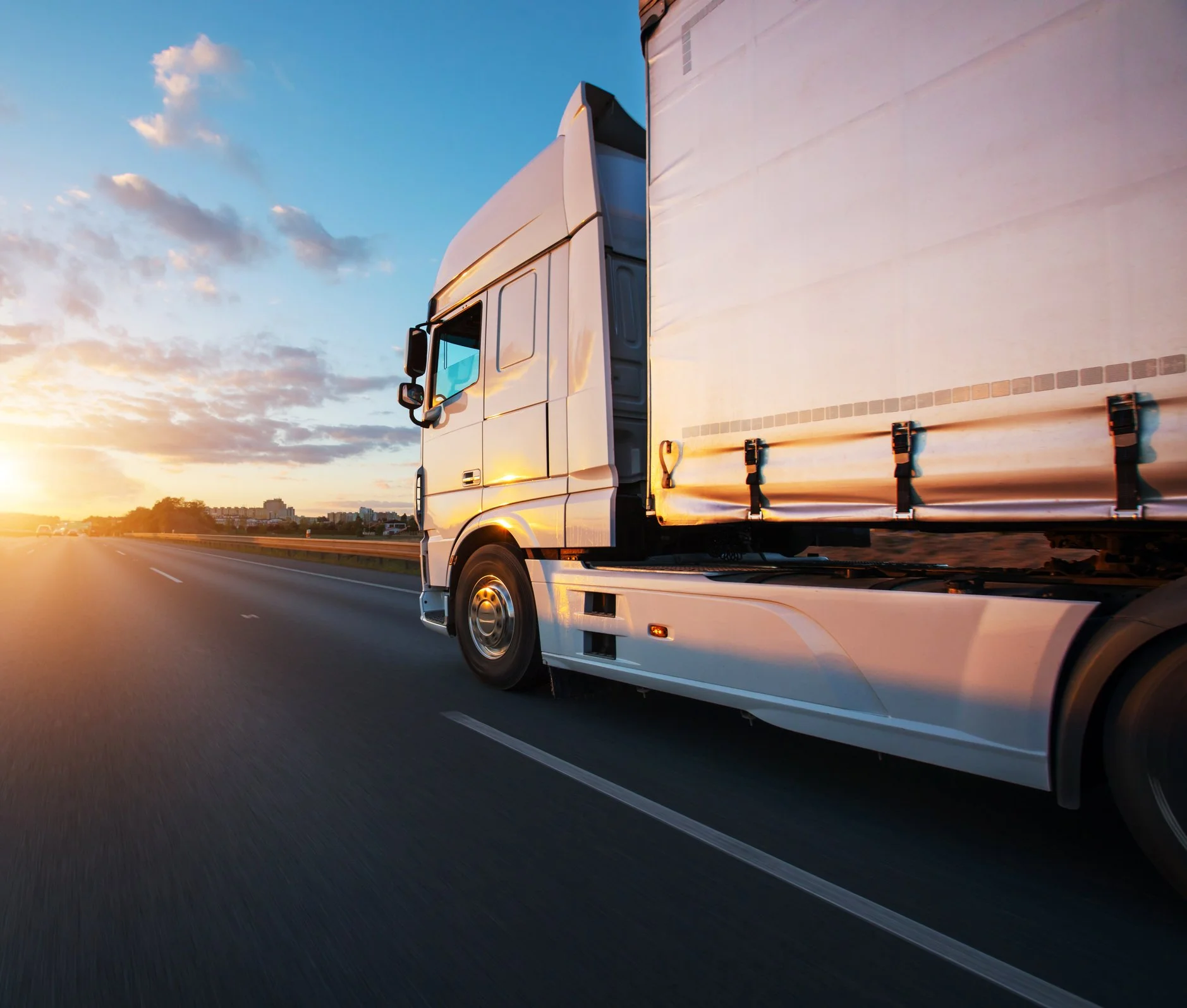 A white semi-truck drives swiftly on a highway at sunset, casting warm hues across the sky.