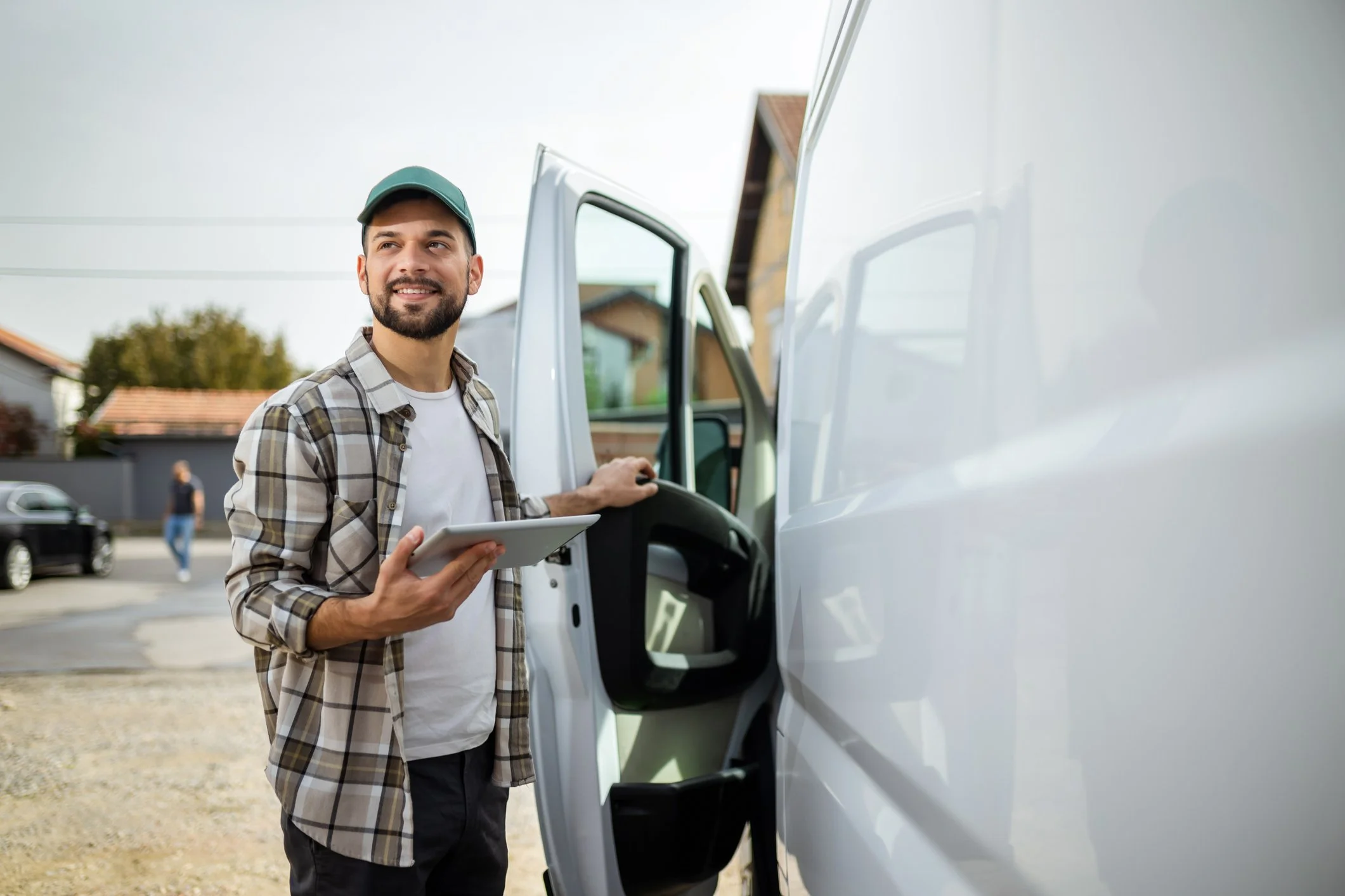 A smiling man in a plaid shirt and cap holds a tablet near an open van door, with a suburban background.