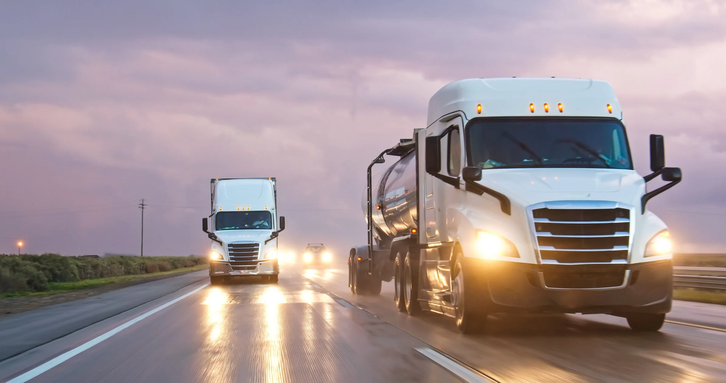 Two white semi-trucks with headlights on traveling on a wet highway under an overcast sky.