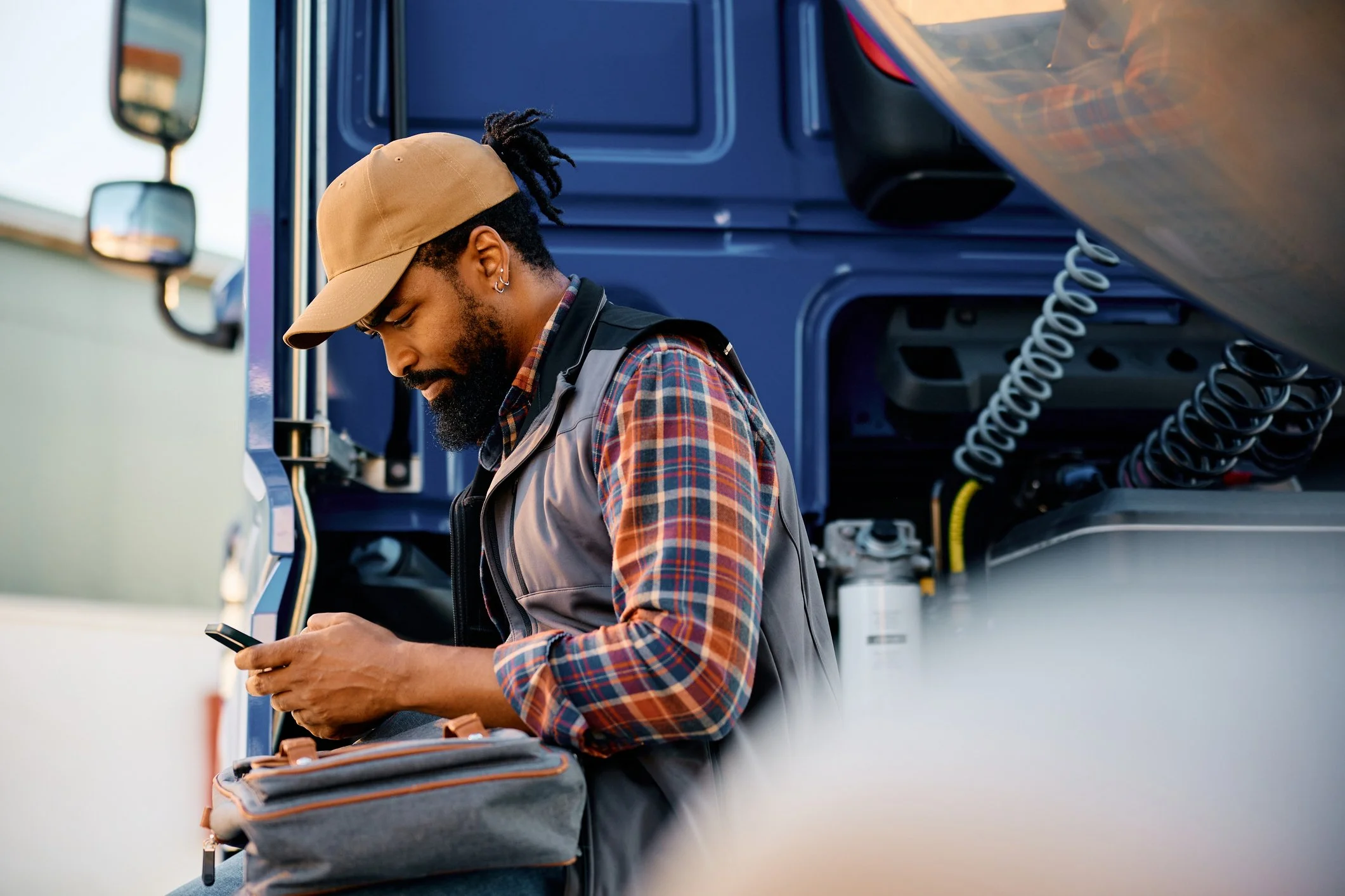 A man in a cap and plaid shirt stands beside a blue truck, intently checking his smartphone.